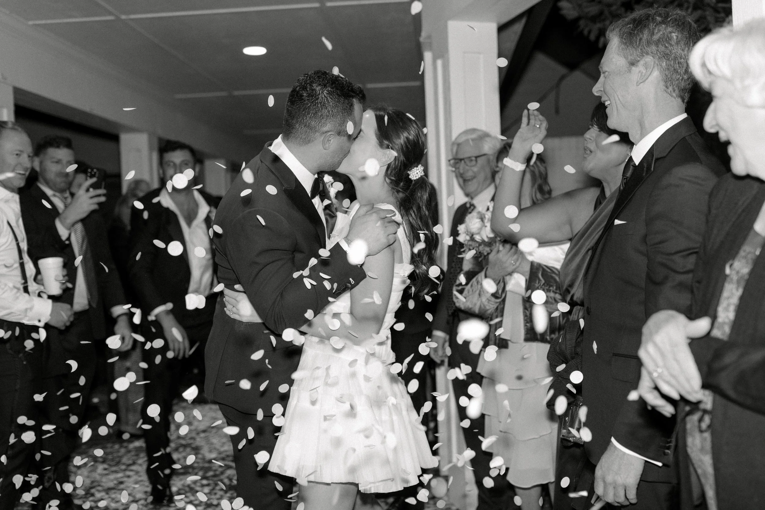 A couple in wedding attire sharing a kiss while surrounded by excited guests throwing confetti, in black and white.