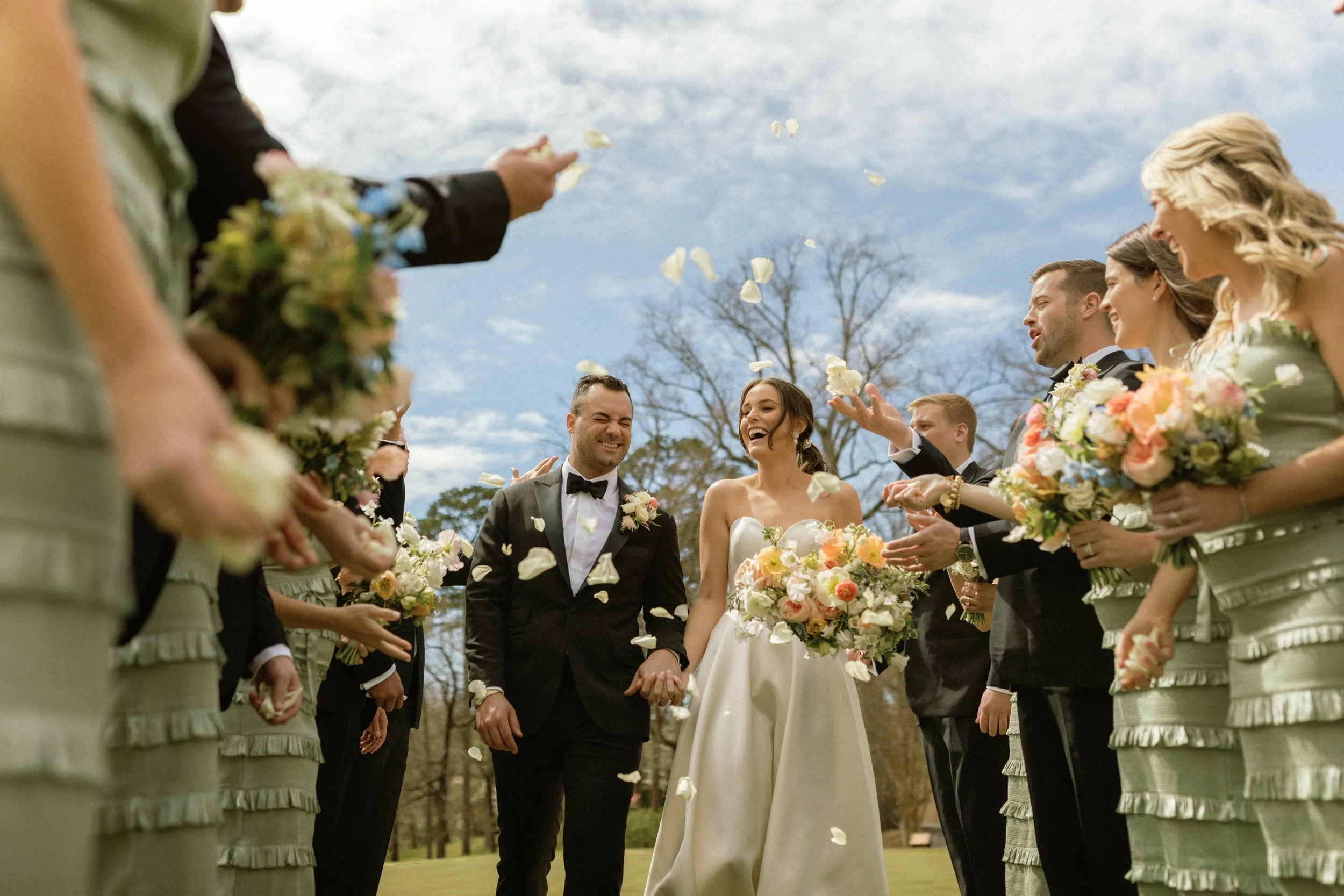 Bride and groom walking hand in hand through a shower of flower petals, surrounded by bridesmaids and groomsmen outdoors on a sunny day.
