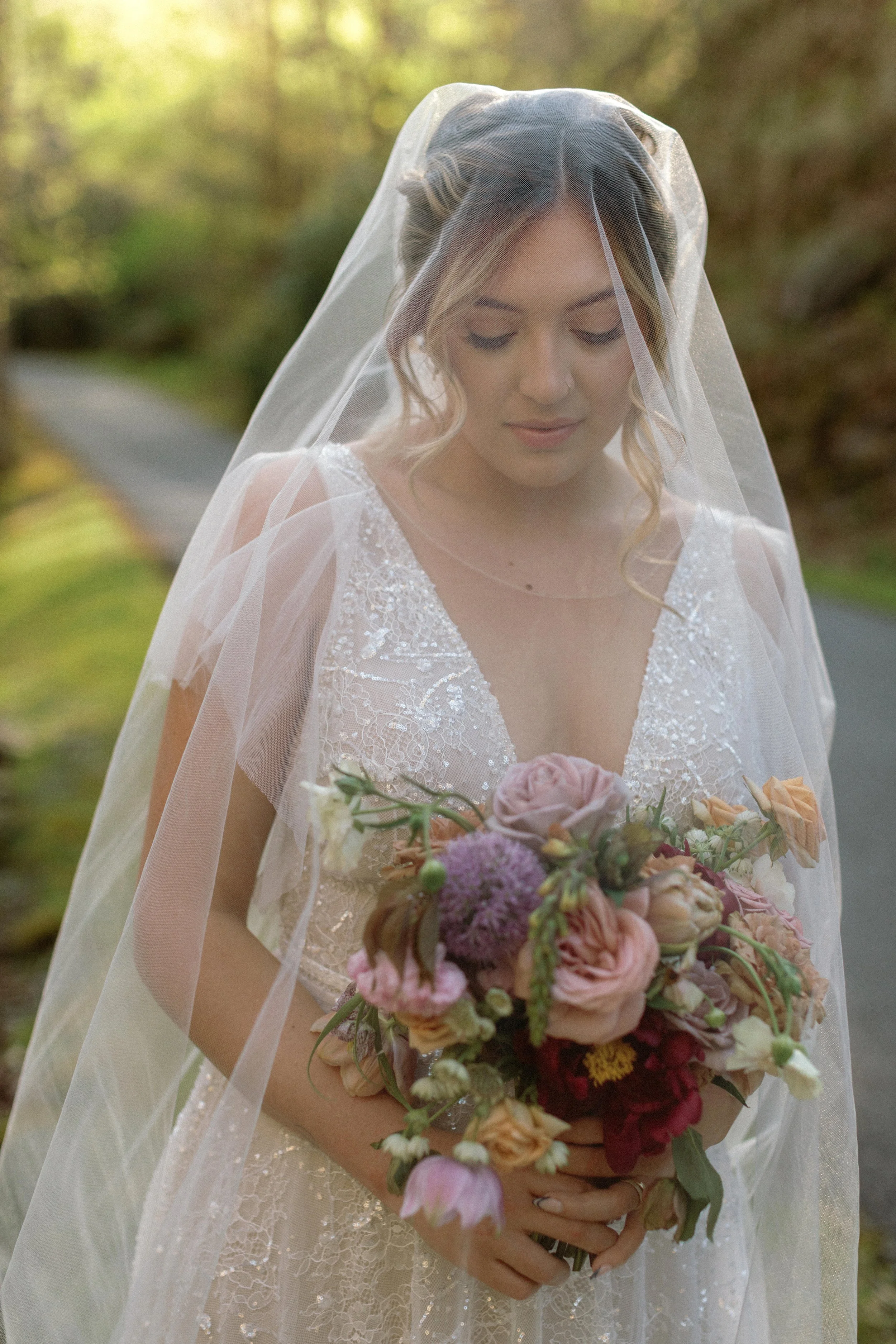 A bride in a lace wedding dress and veil holding a bouquet of pink, purple, and peach flowers, standing outdoors on a path surrounded by greenery.