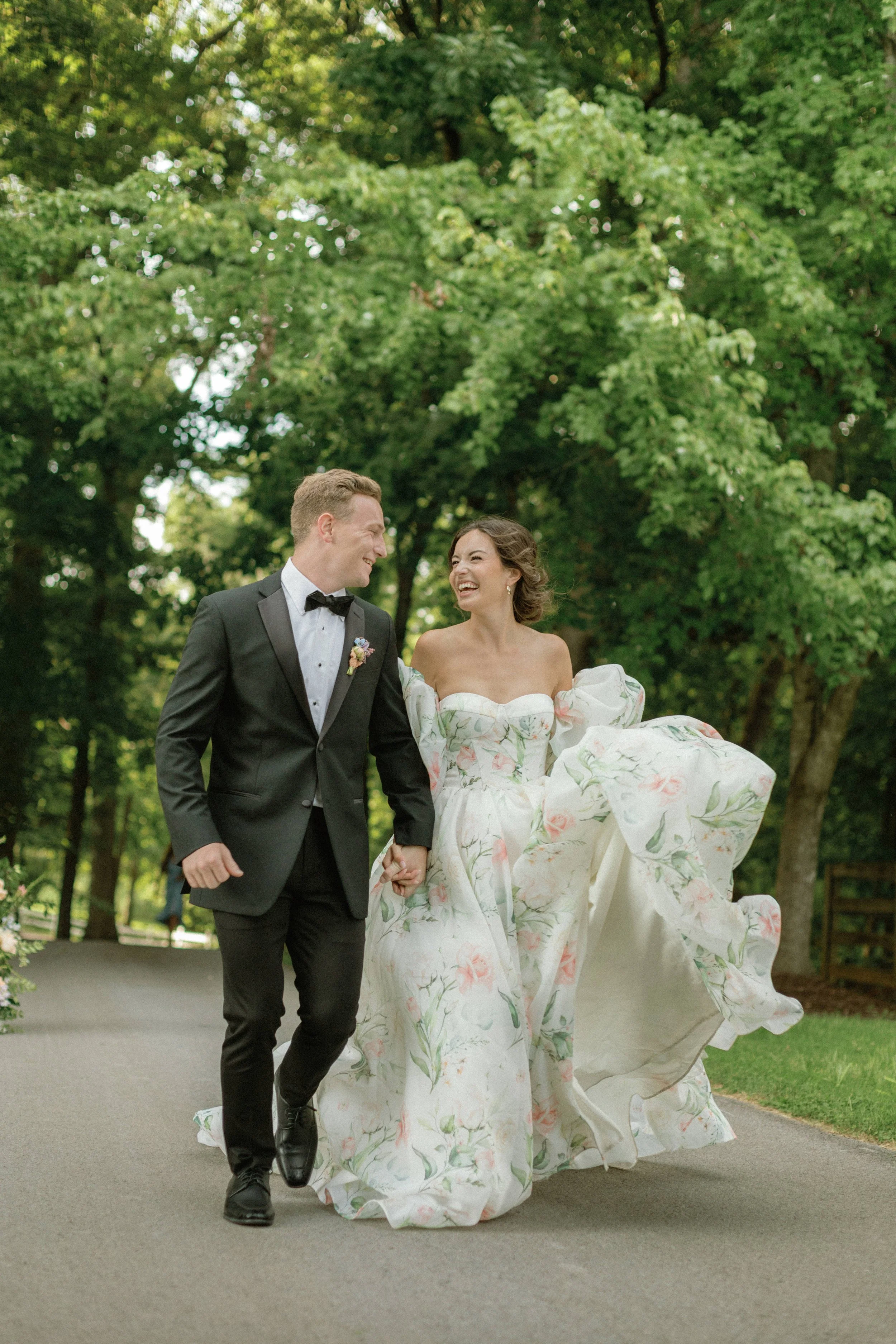 A newlywed couple walking hand in hand outdoors on a paved path, smiling and laughing. The bride is wearing a white wedding gown with pink and green floral patterns and puffed sleeves, and the groom is in a black tuxedo with a bow tie. Green trees su