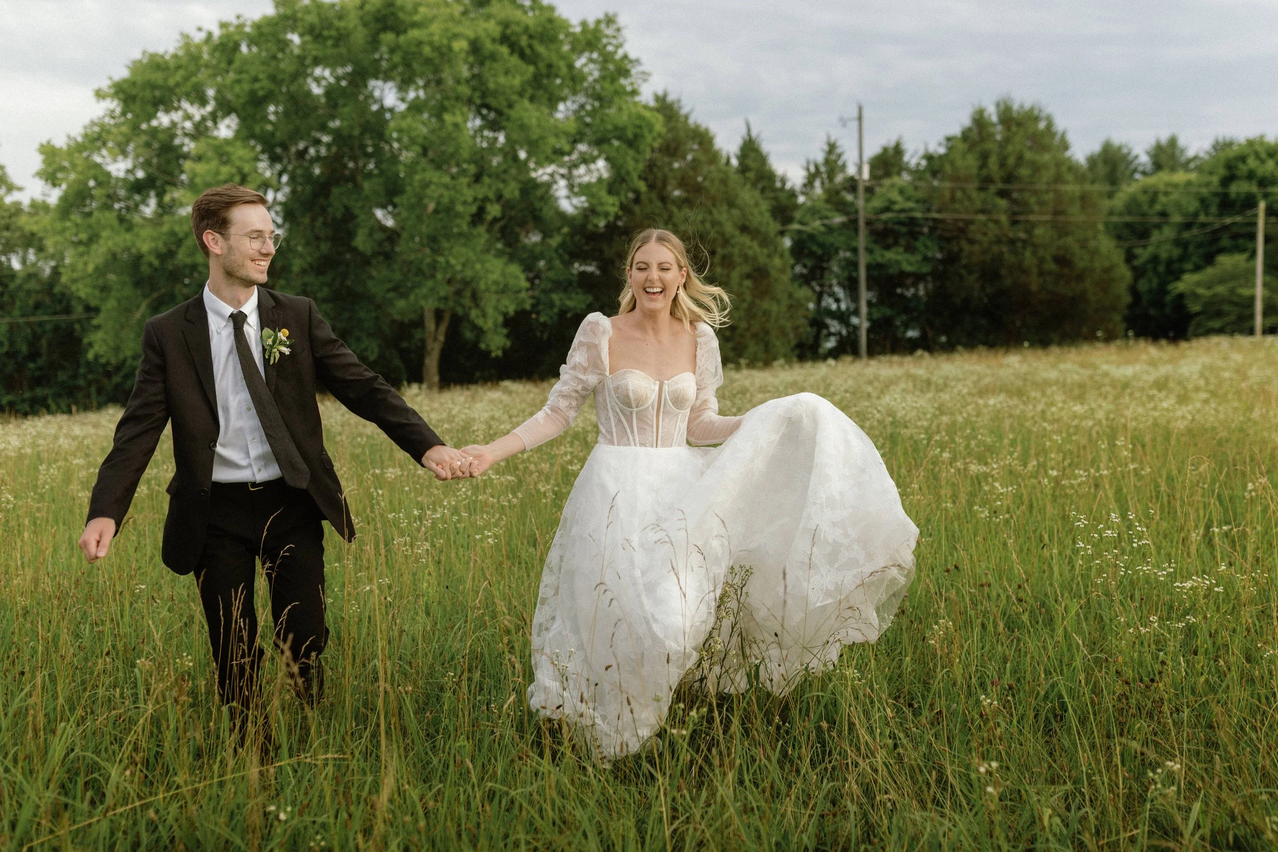 A couple holding hands and smiling, walking through a grassy field on their wedding day, with lush green trees and cloudy sky in the background.