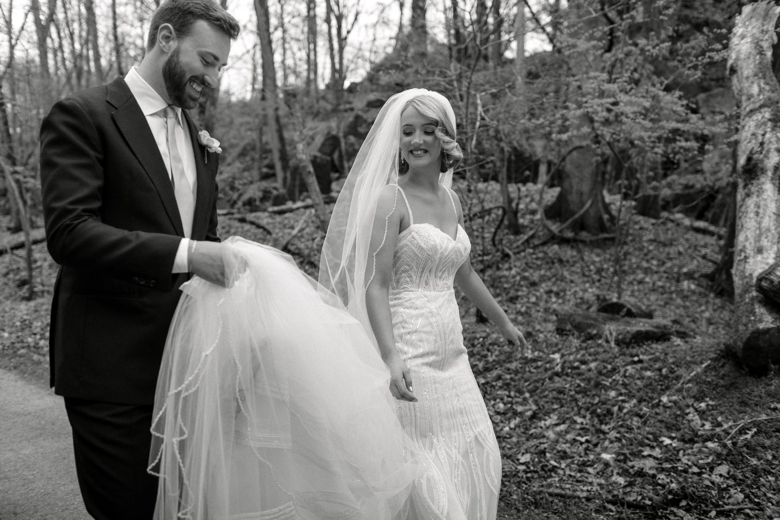A black and white photo of a groom lifting the bride's wedding dress, both smiling, in a wooded area during their wedding.