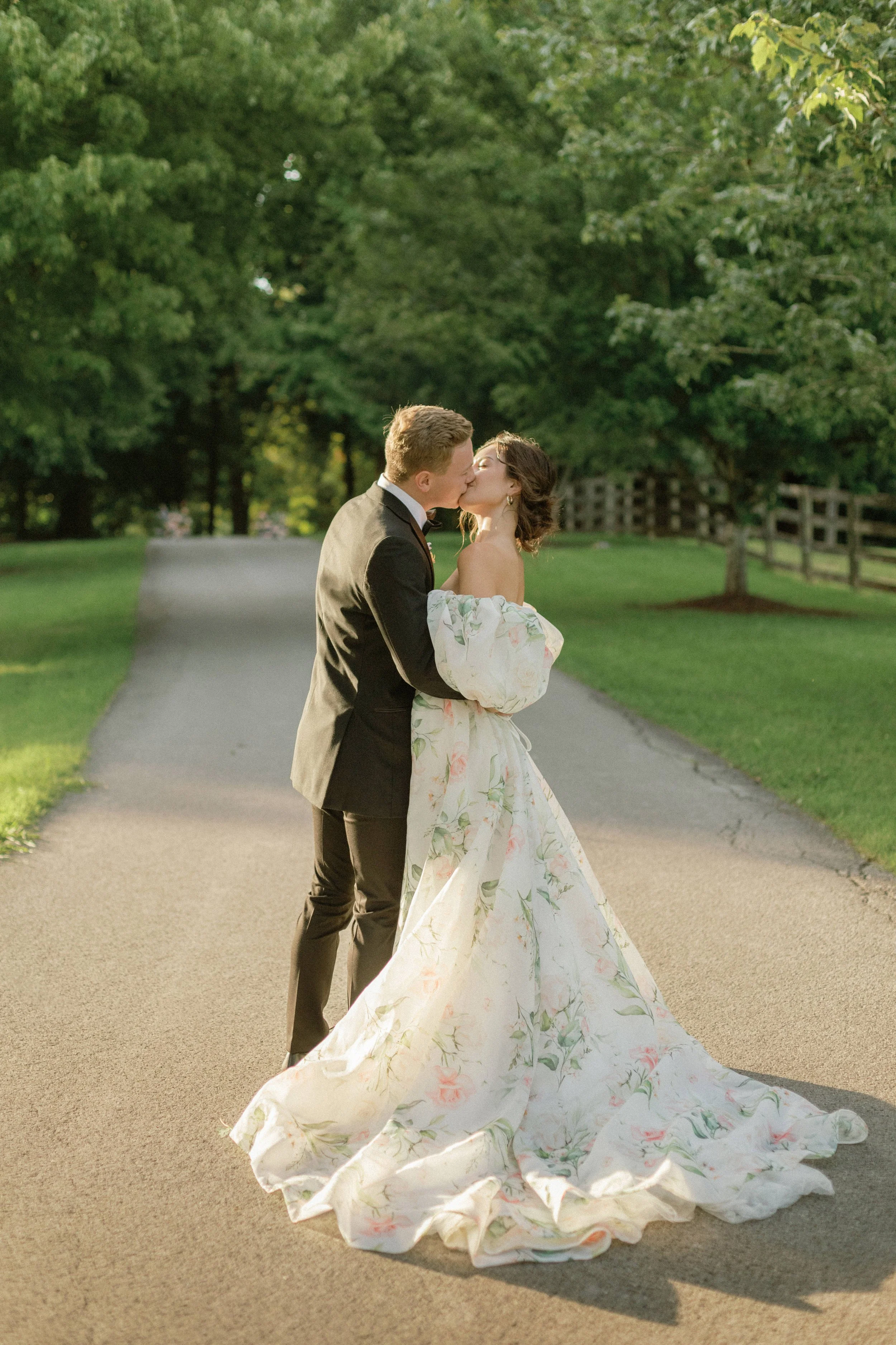 A newly married couple sharing a kiss outdoors on a tree-lined path, with the bride wearing a flowing floral wedding gown and the groom in a black tuxedo.