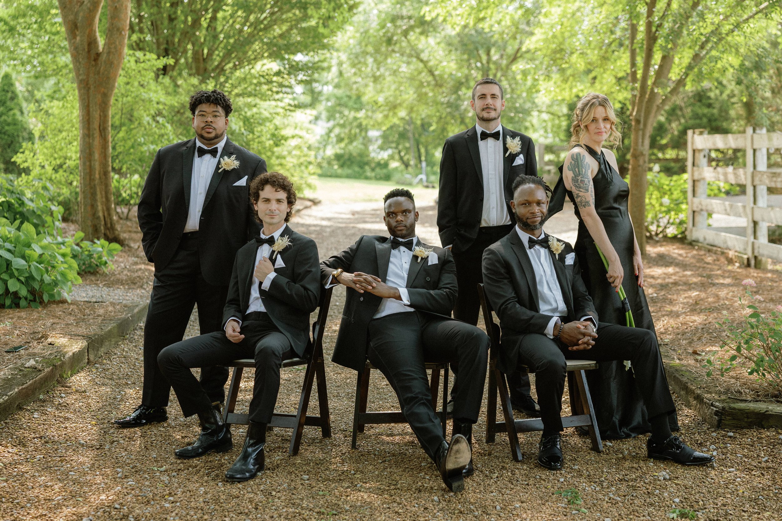 Group of six diverse people dressed in formal attire outdoors, with trees and greenery in the background, posing for a photo.