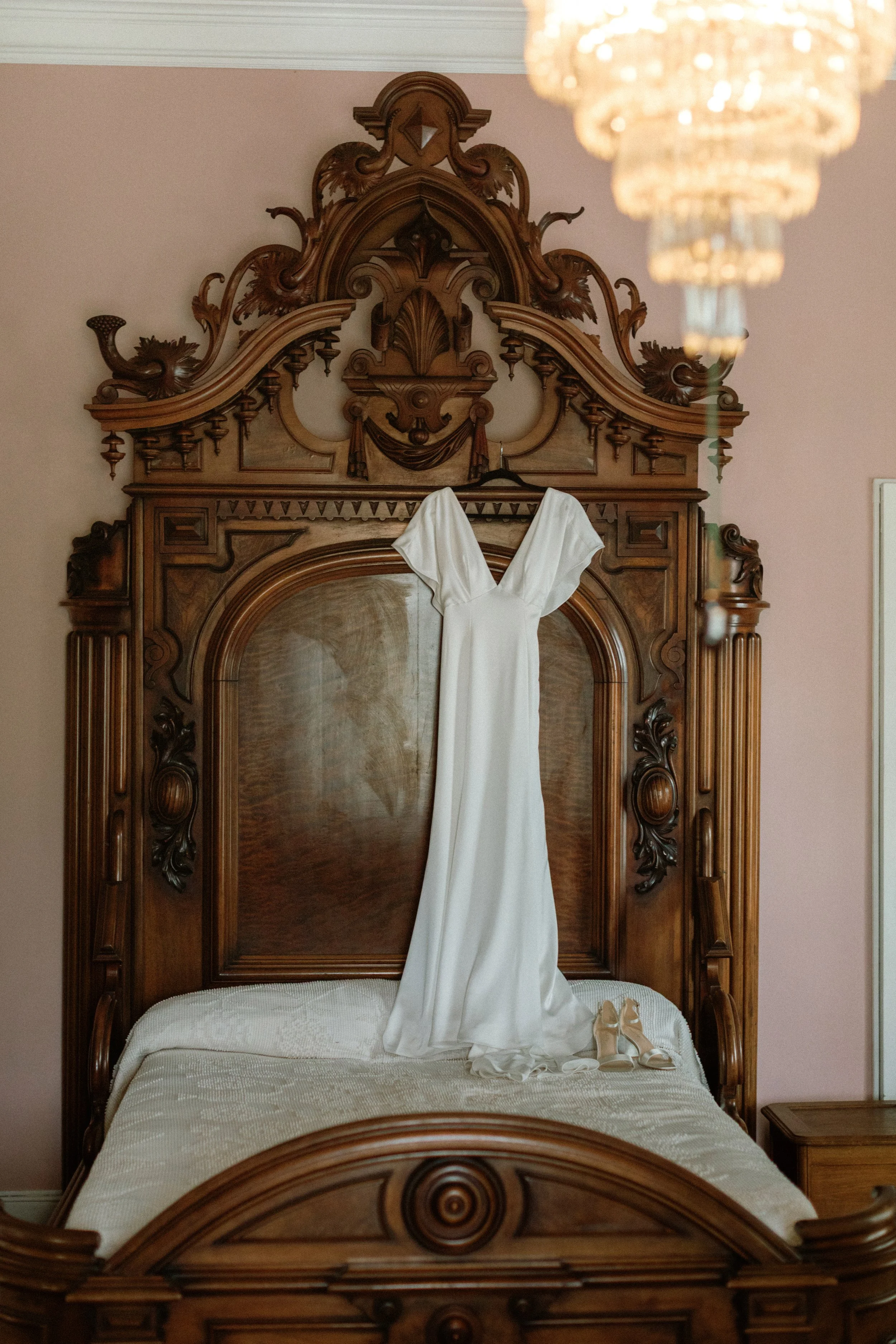 A white wedding dress hanging on a vintage wooden bed headboard, with a pair of beige high heels placed on the bed.
