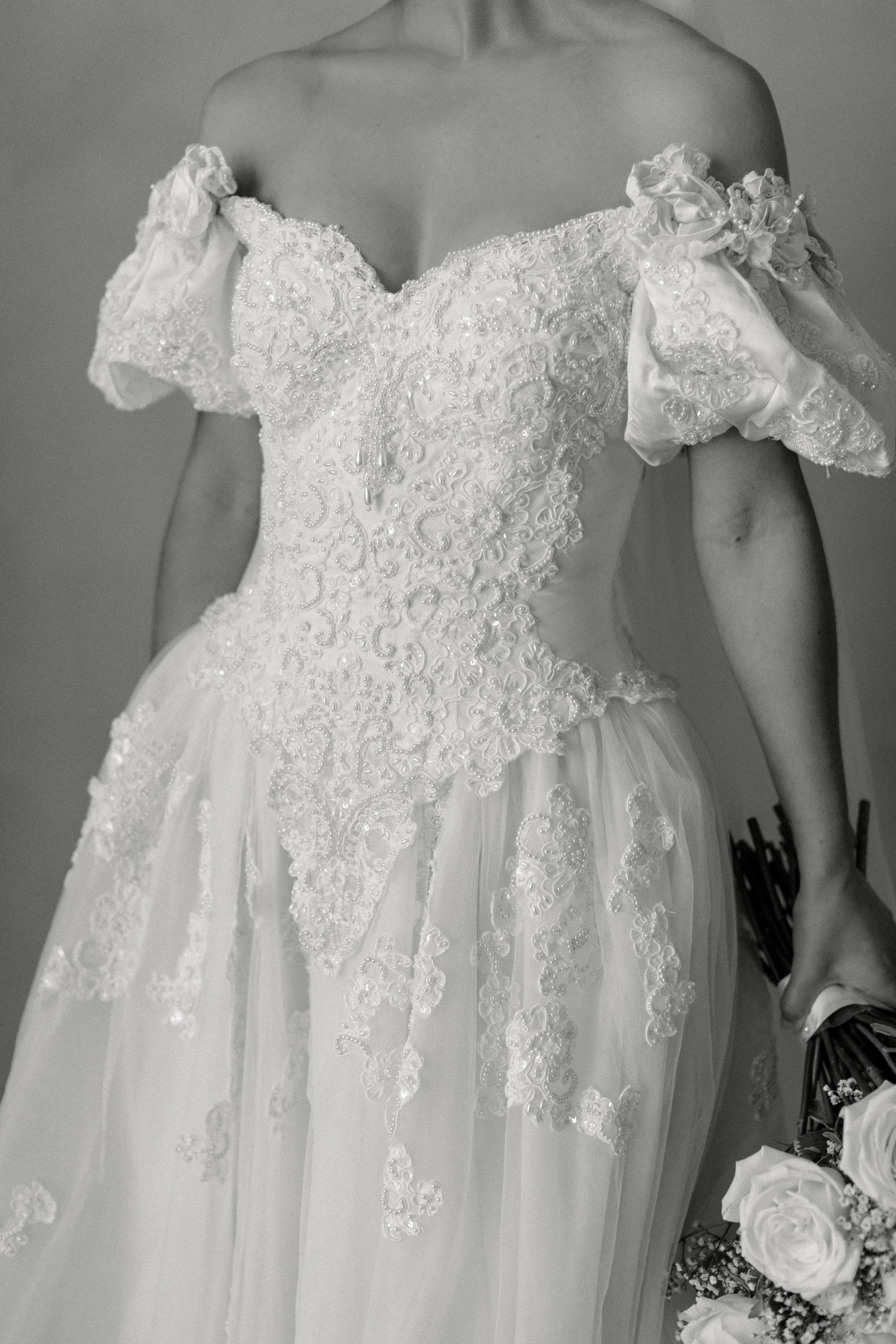 A woman wearing an ornate, white wedding dress with lace details and off-the-shoulder puff sleeves, holding a bouquet of roses in her right hand.