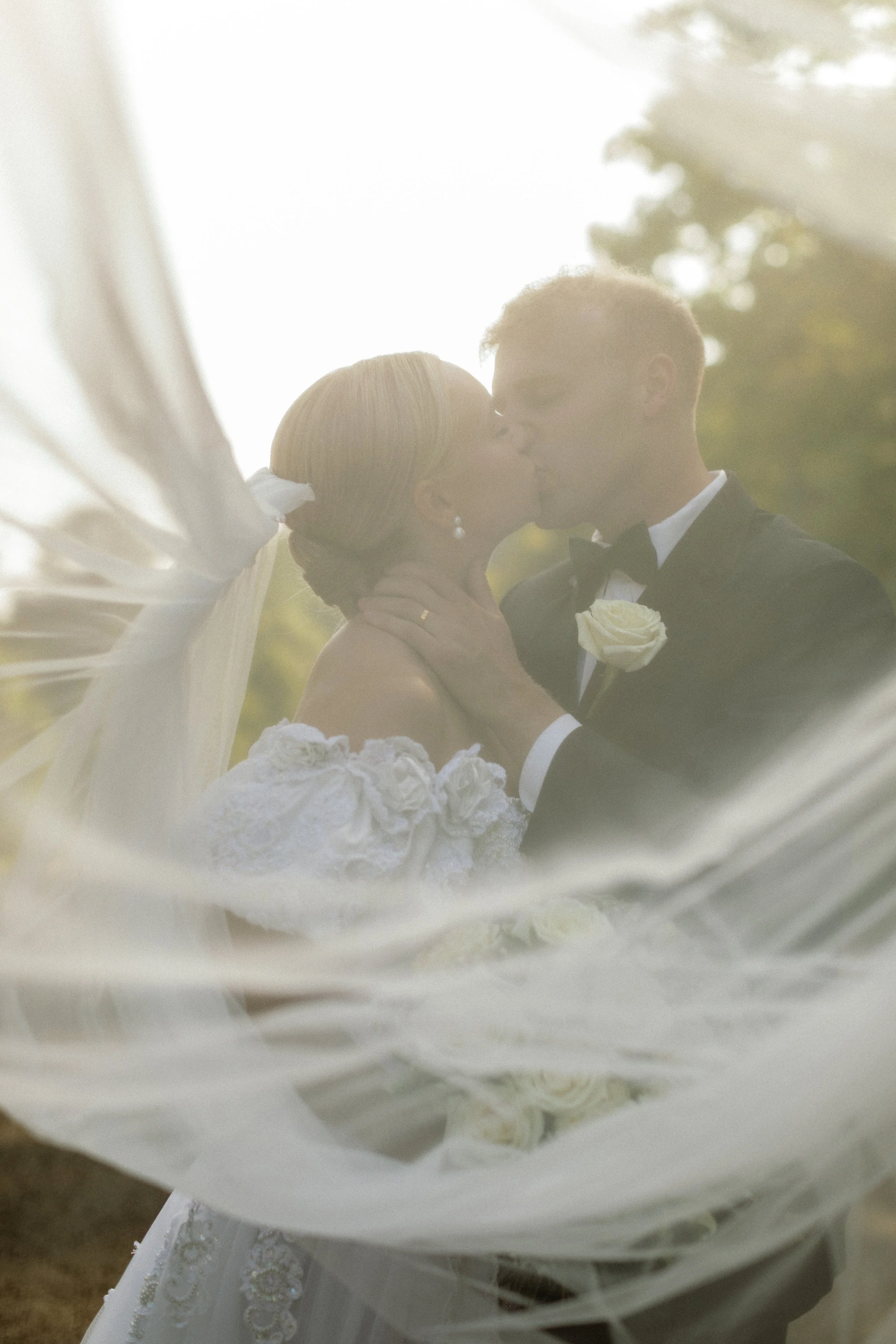 A couple sharing a kiss on their wedding day, framed through a delicate veil with soft sunlight behind them.