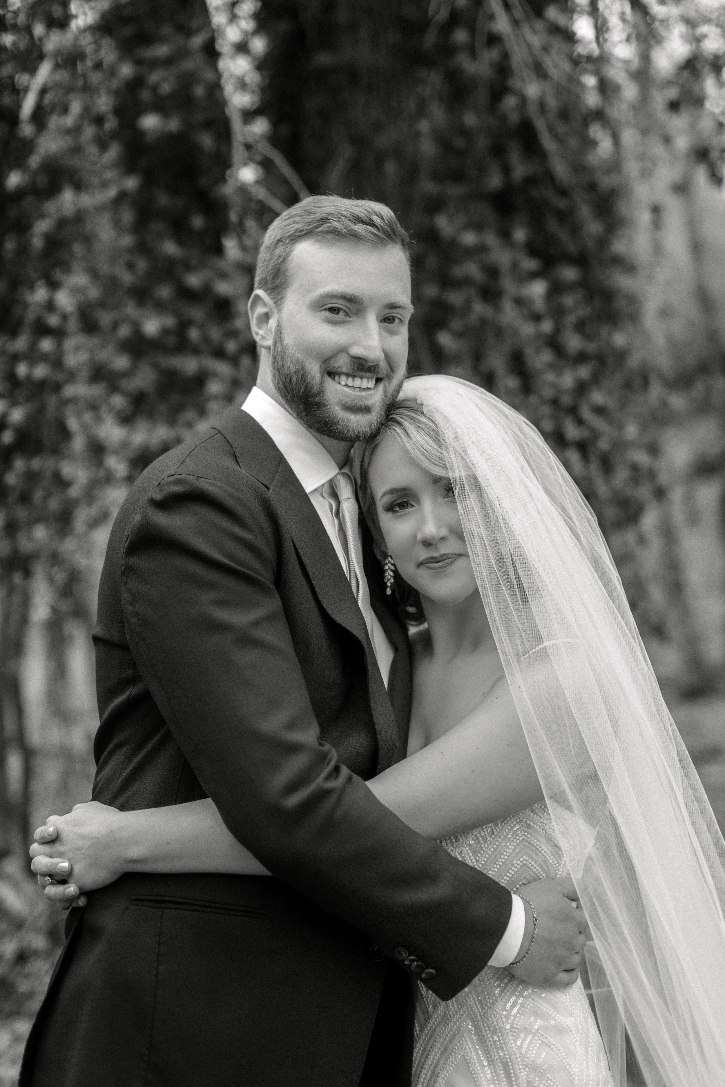 Black and white photograph of a smiling groom and a bride with a veil hugging outdoors in front of trees.