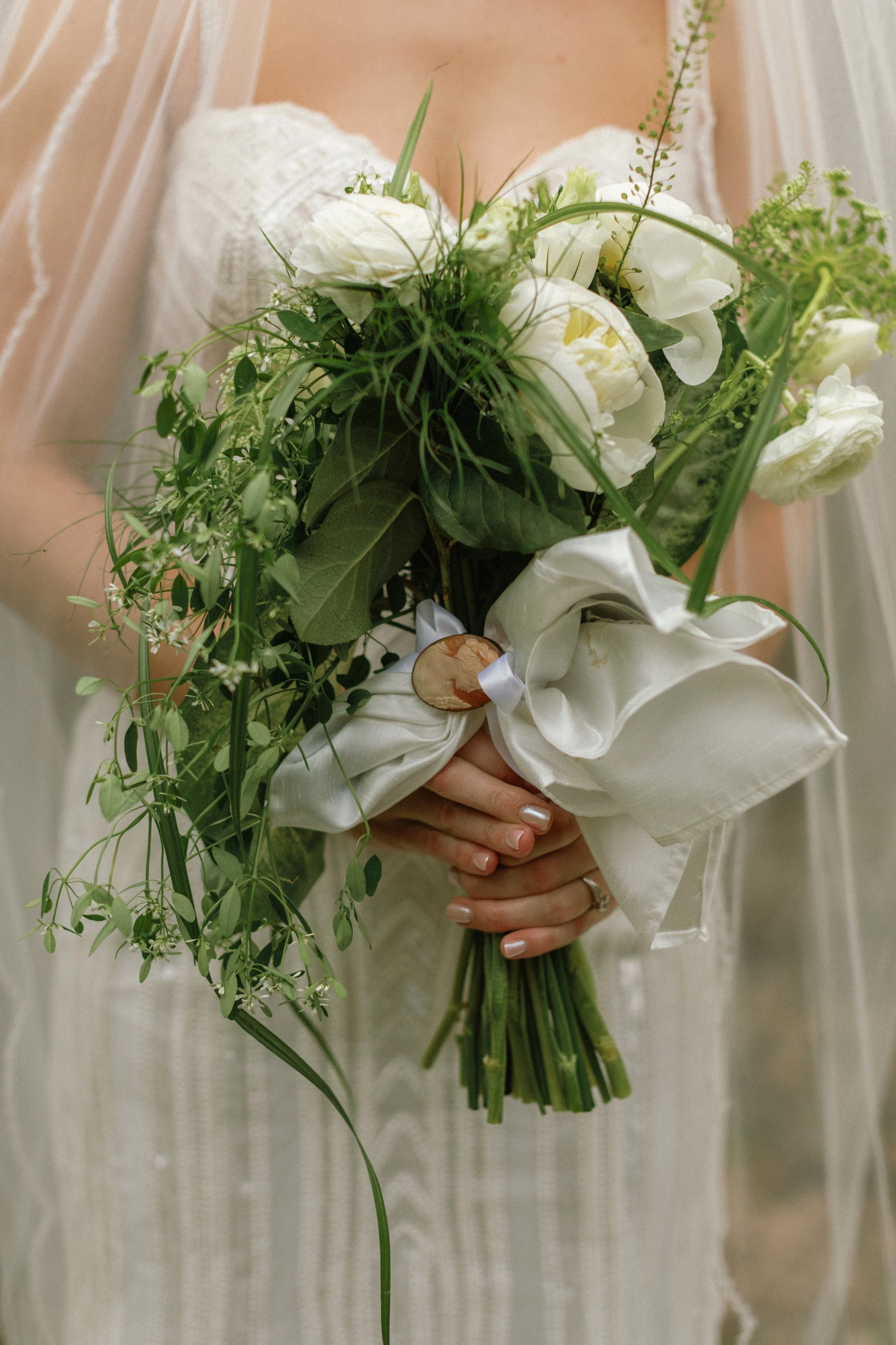 Close-up of a woman in a white wedding dress holding a bouquet of white flowers and greenery, wrapped with white fabric and a decorative button.