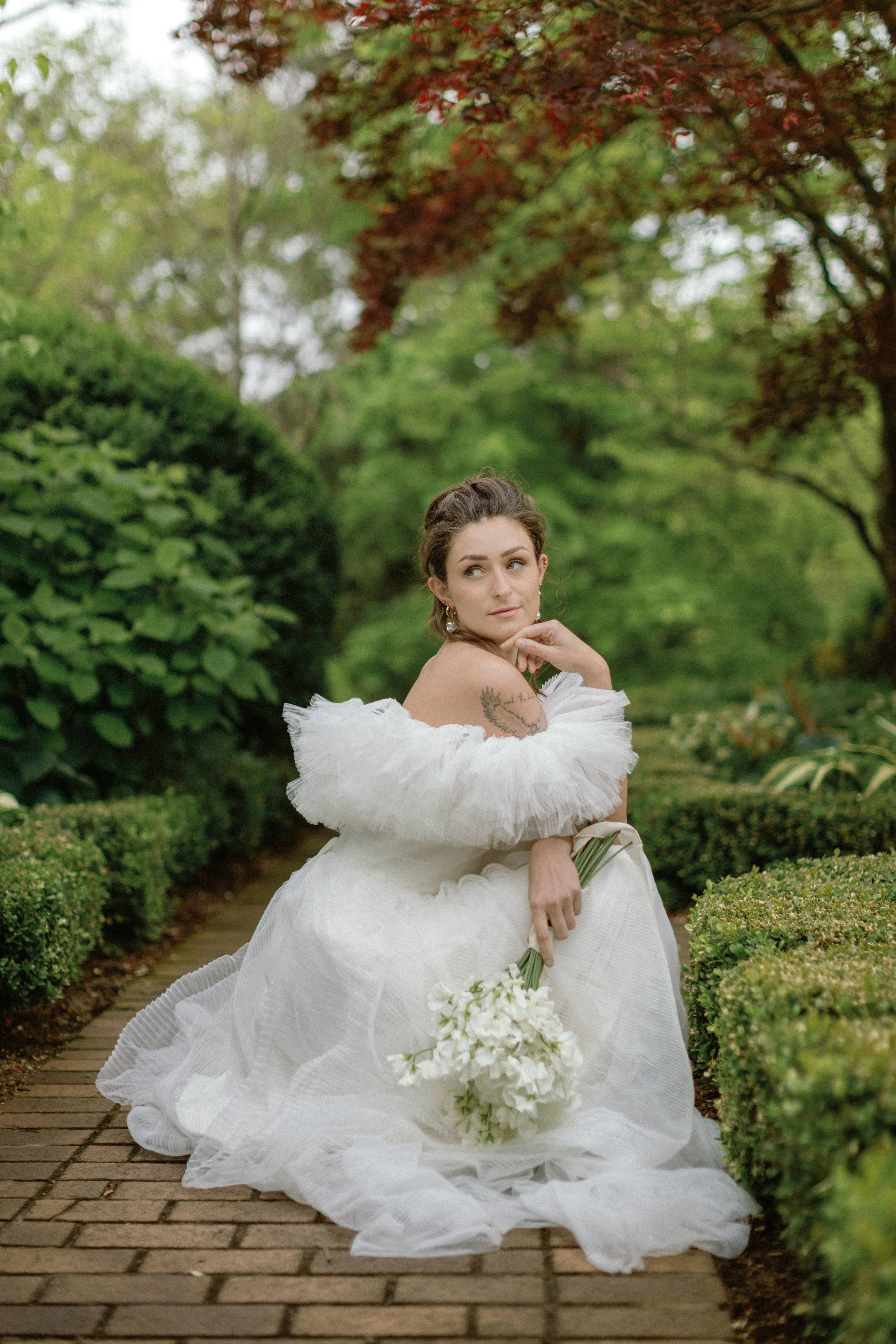 Woman in a white wedding dress sitting on a brick pathway in a garden, holding a bouquet of white flowers, surrounded by lush greenery.