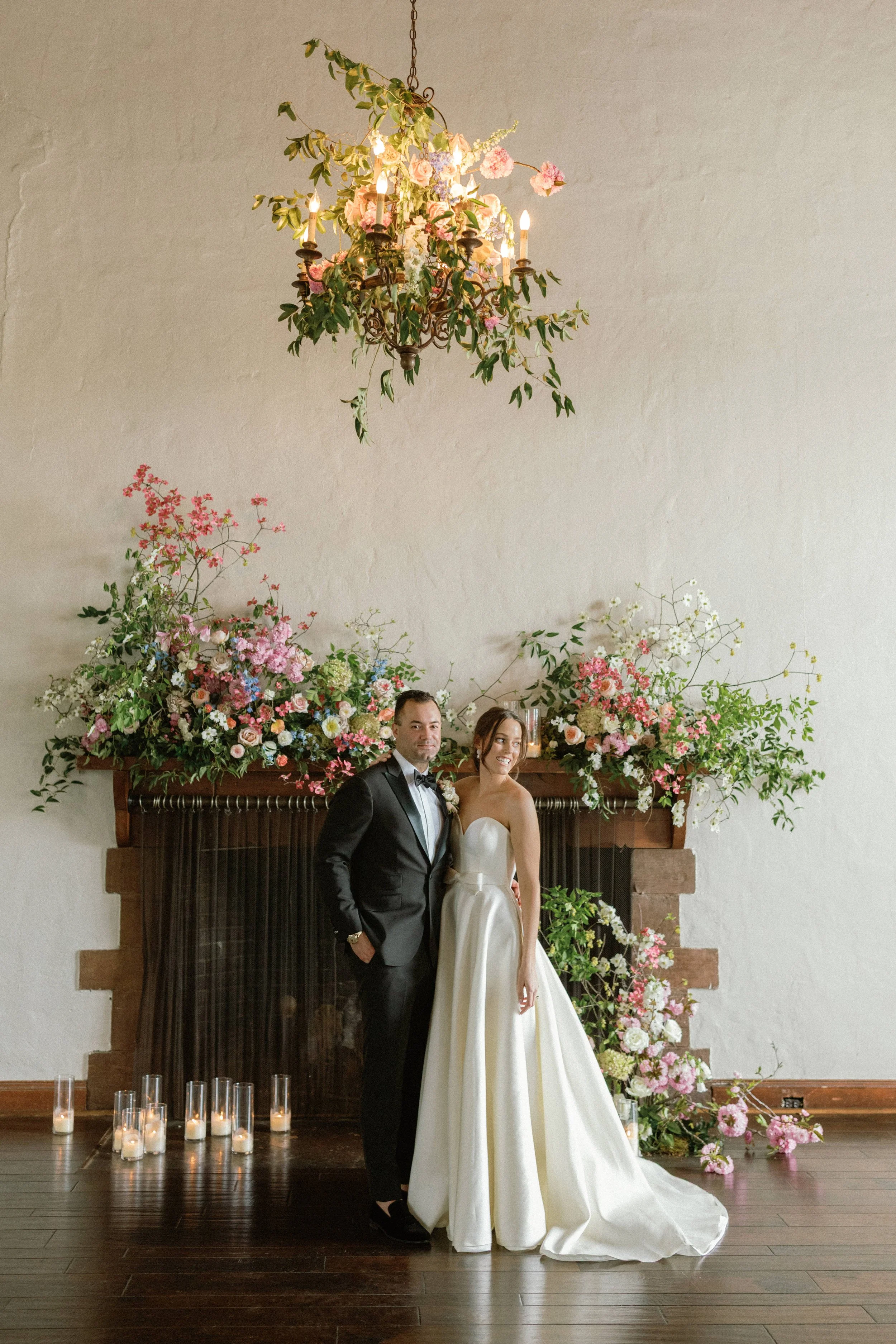 A bride and groom in wedding attire standing together in front of a floral arrangement on a fireplace mantel, with a chandelier hanging above and candles on the floor.