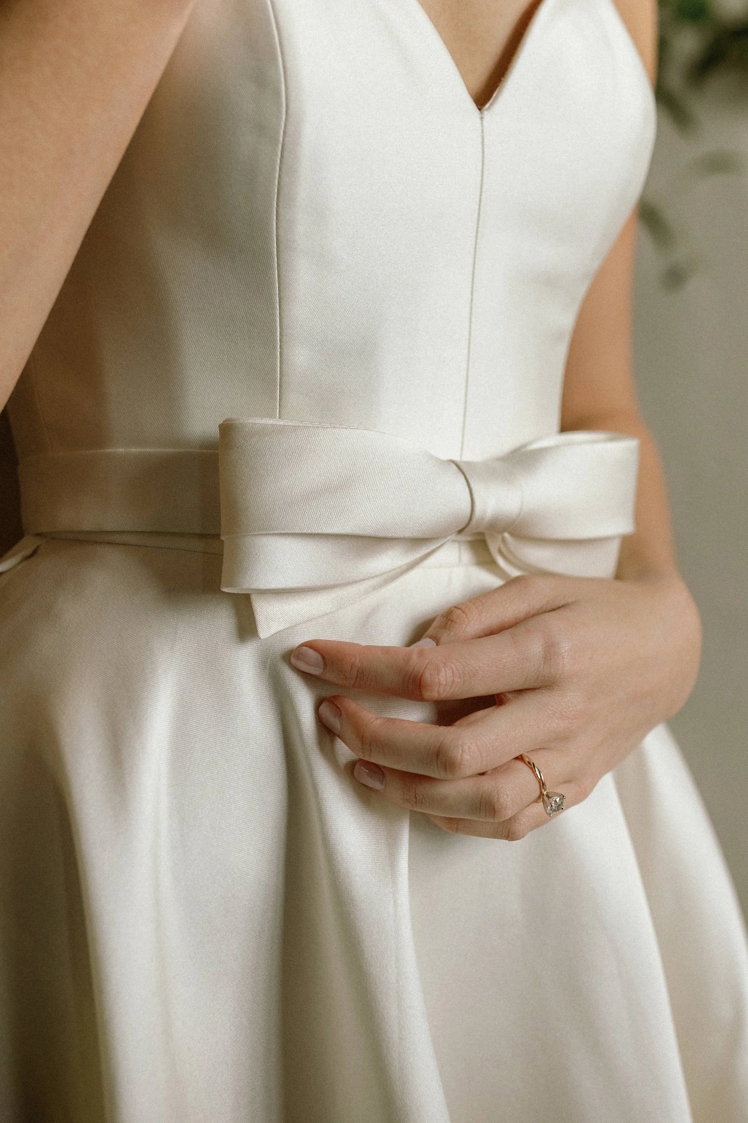 Close-up of a woman wearing a white dress with a bow at the waist, her hand resting on her stomach, showing a ring on her finger.
