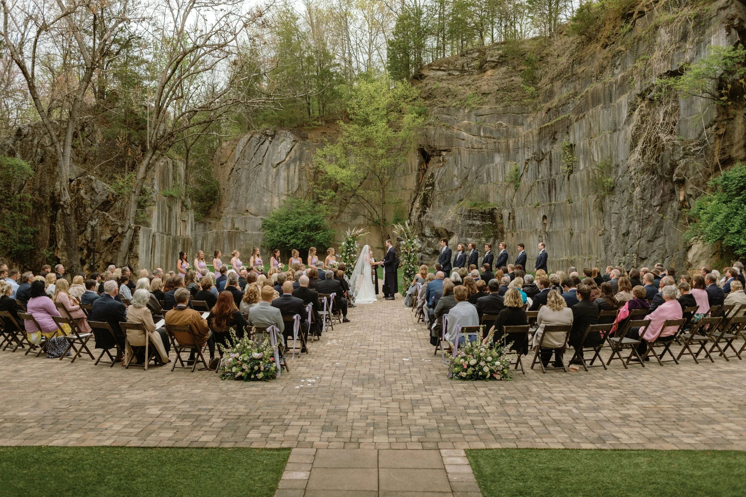 Outdoor wedding ceremony taking place in front of a rocky cliff with green trees, where a bride and groom are exchanging vows. Bridesmaids and groomsmen are standing on either side, and guests are seated in rows on a brick pathway.