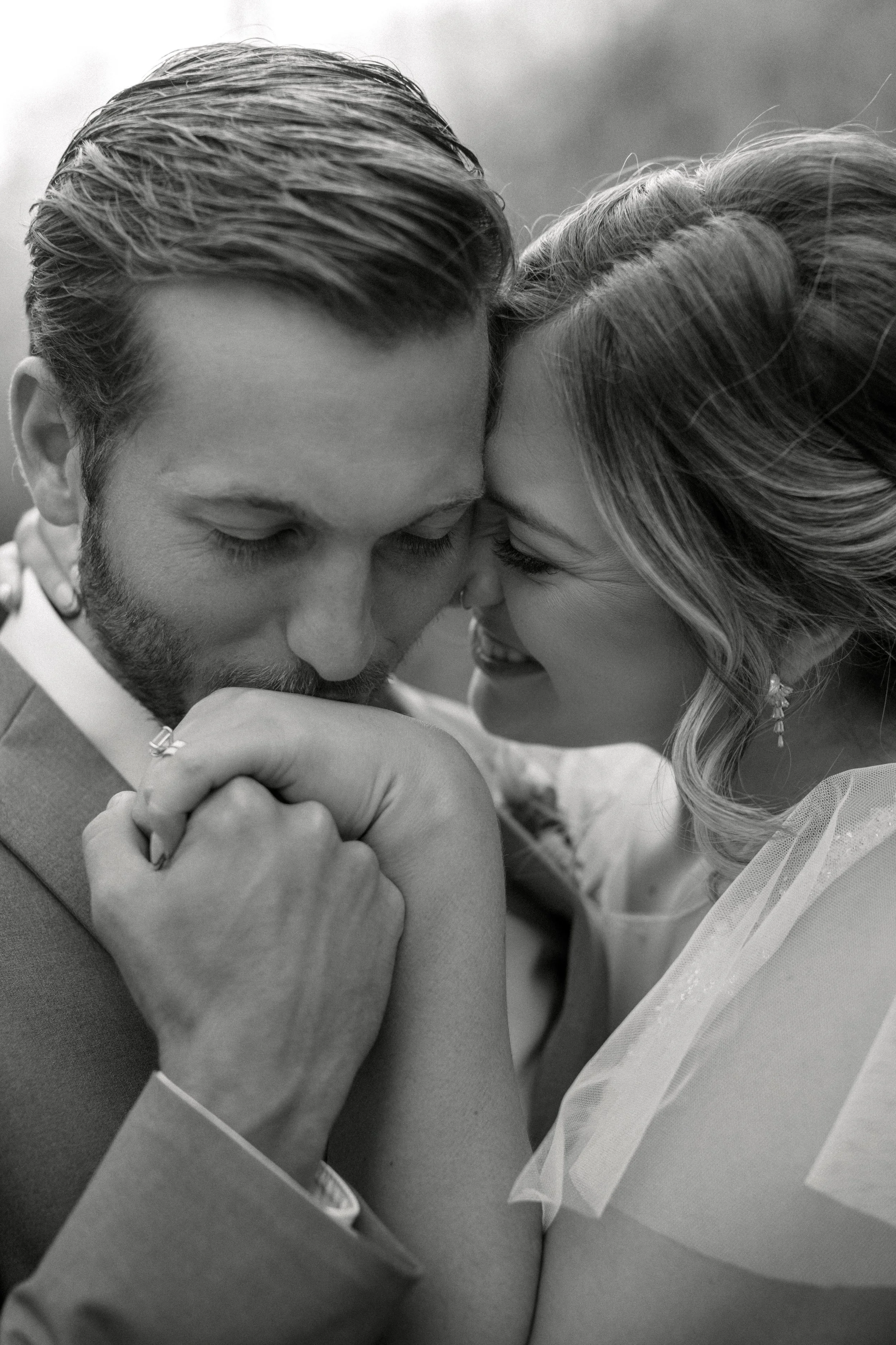 A black and white close-up photo of a bride and groom with their foreheads touching, holding hands, smiling, and sharing an intimate moment during their wedding ceremony.
