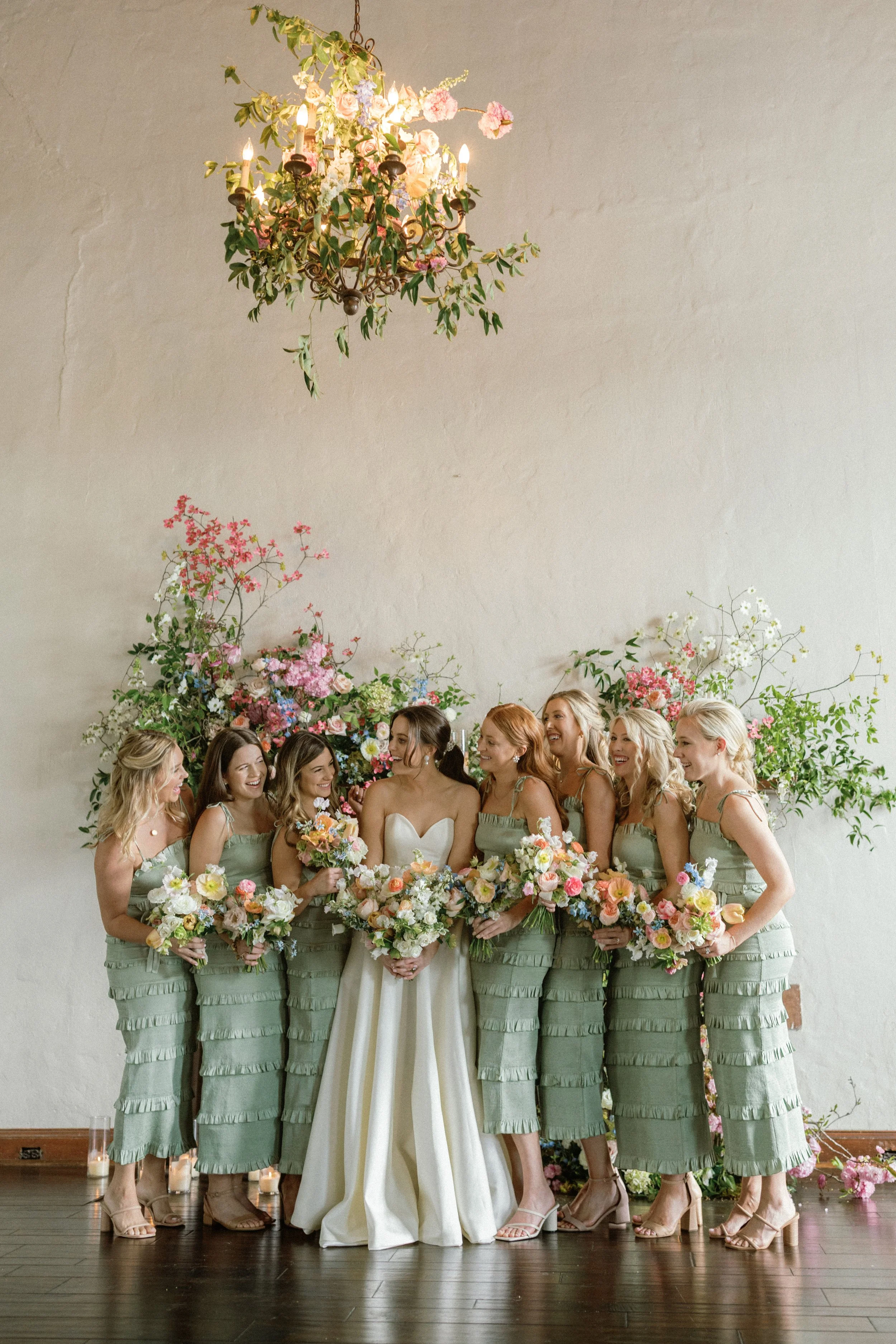 A bride and her seven bridesmaids standing together at a wedding, holding bouquets of flowers, with floral arrangements behind them and a chandelier above.