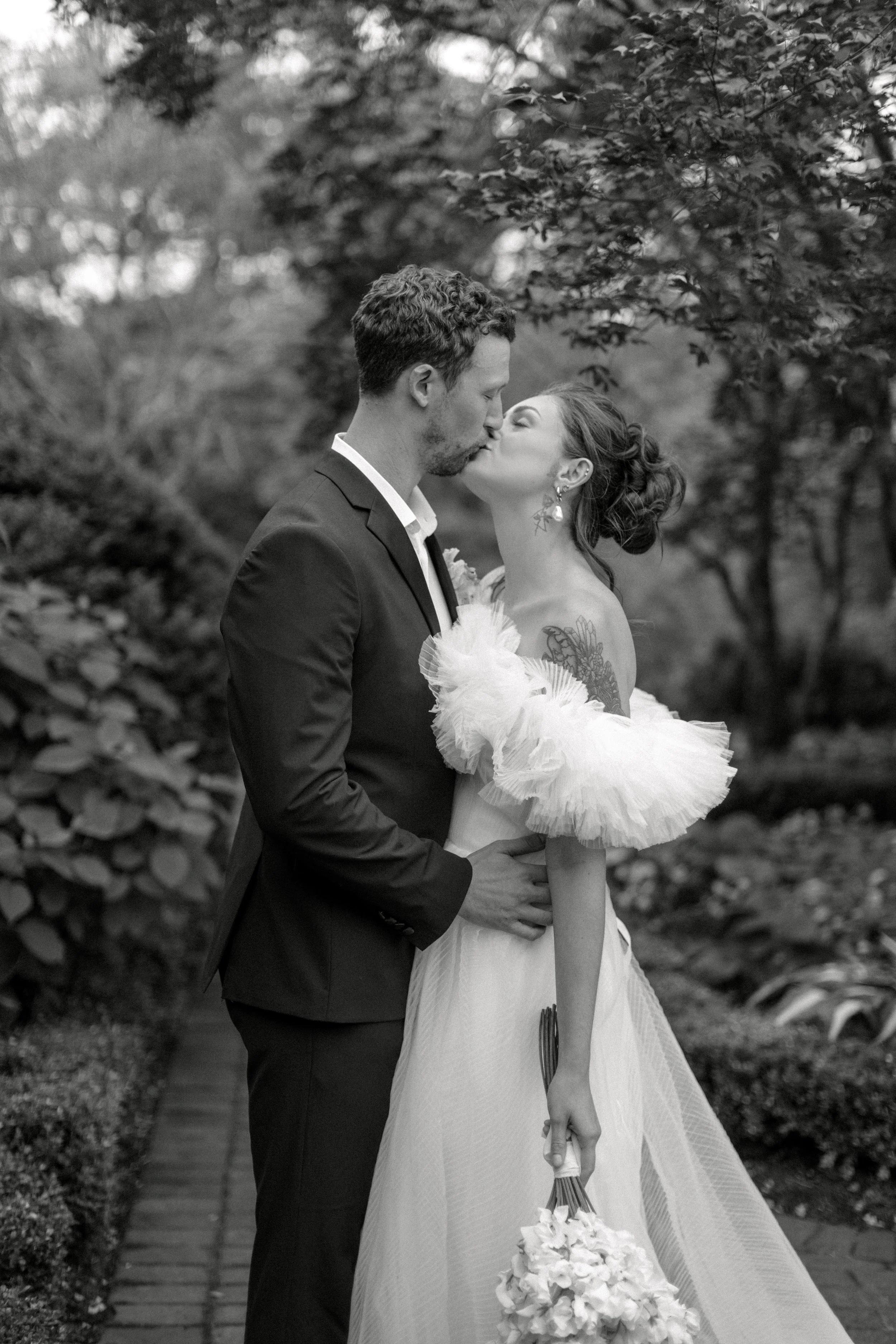 A black and white photo of a bride and groom sharing a kiss outdoors, the bride holding a bouquet and wearing an off-shoulder dress with ruffled sleeves, and the groom in a suit surrounded by trees and foliage.