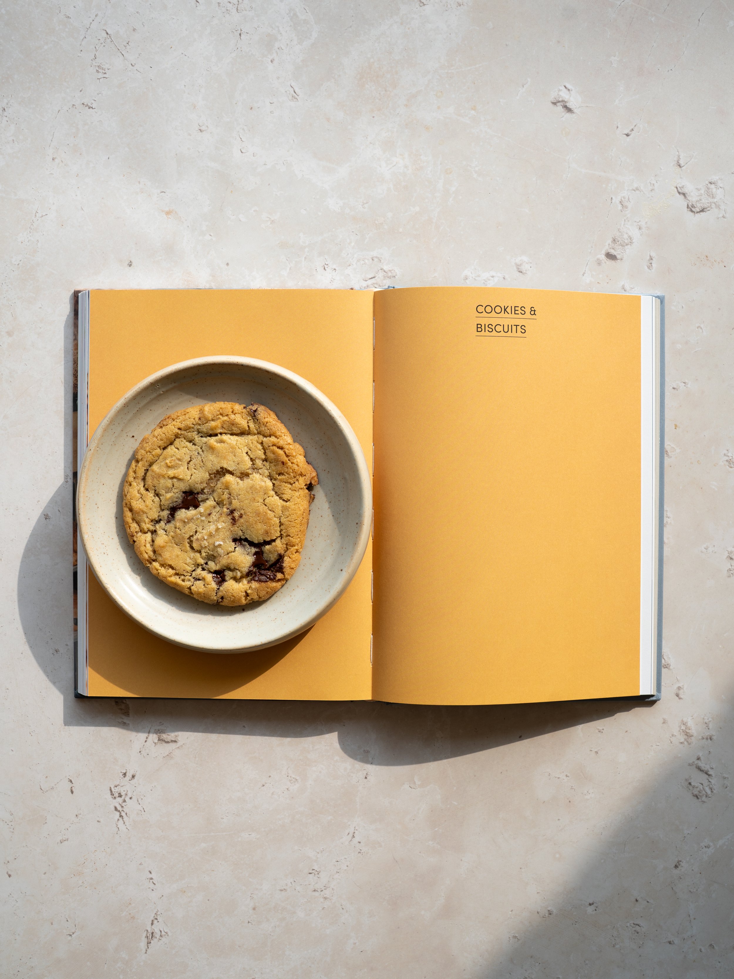 Open notebook with yellow pages and the words "Cookies & Biscuits" on the right page, with a plate containing a chocolate chip cookie on the left page, on a light-colored surface.