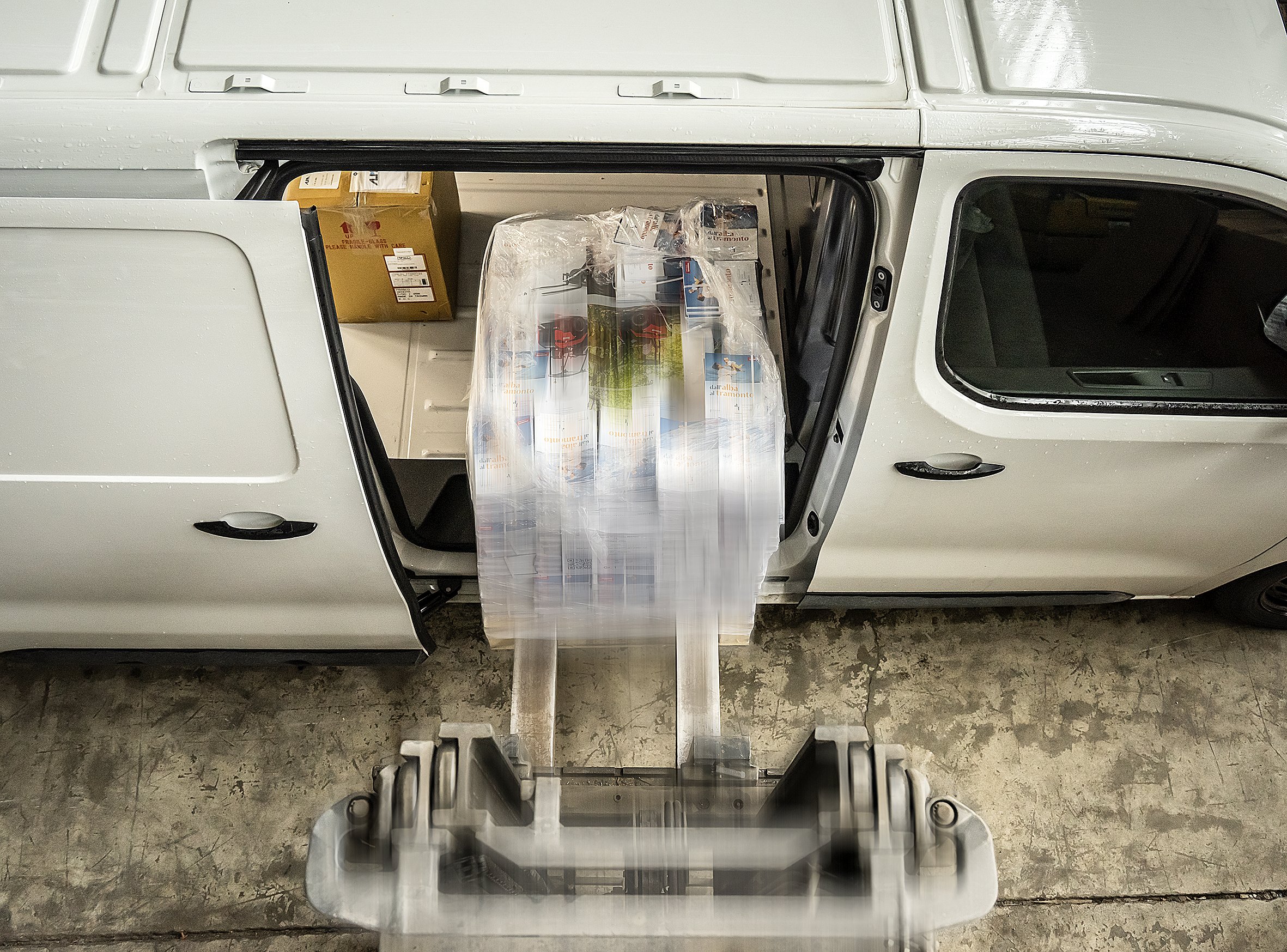 Un furgone bianco con il portellone laterale aperto. All'interno, un pallet di scatole impilate e avvolte in pellicola trasparente viene caricato con un carrello elevatore. Si intravede un altro scatolone nel vano.