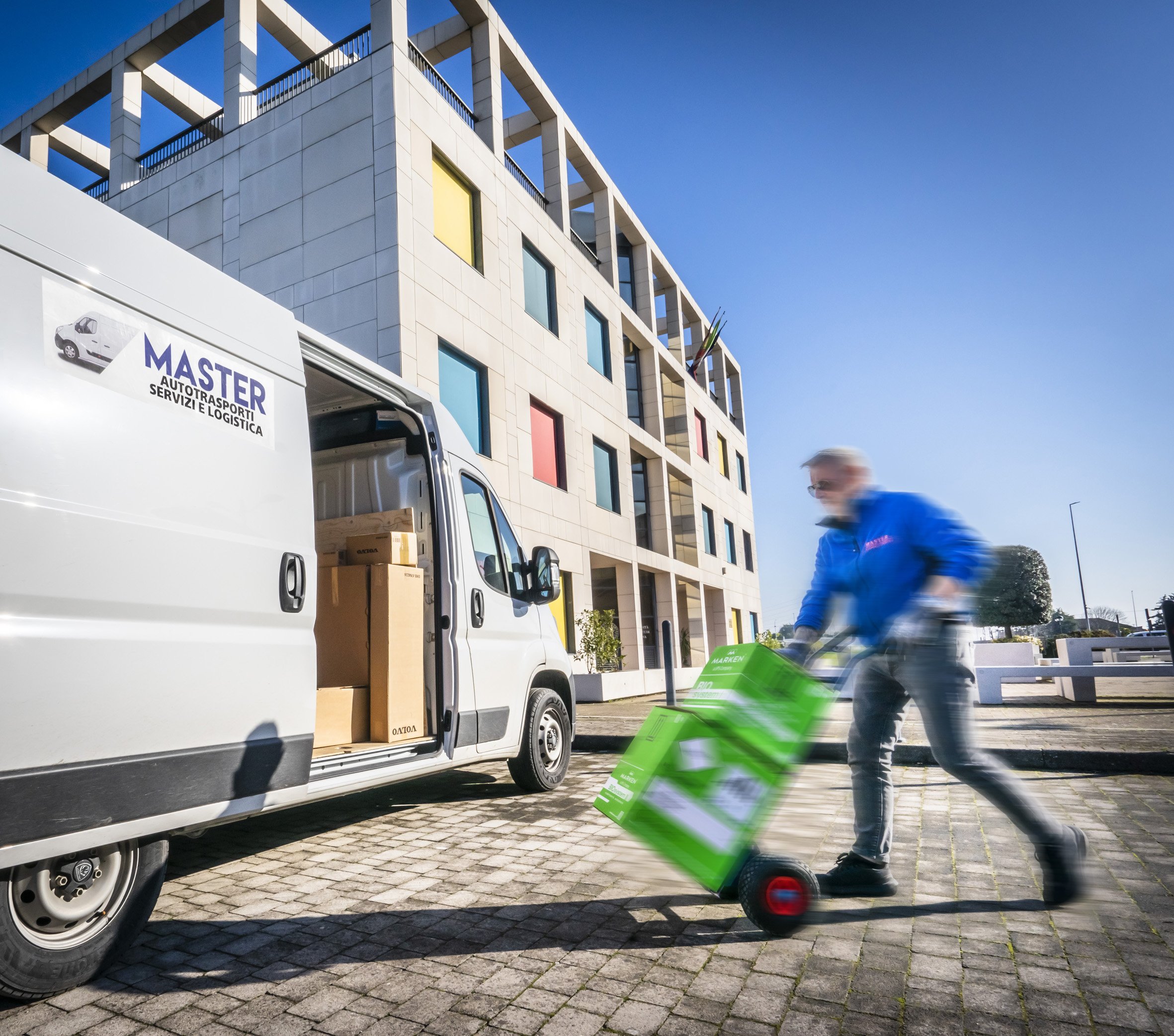 Furgone bianco per trasporti con scritte "Master Autotrasporti Servizi Logistica" parcheggiato con portellone aperto e scatole all'interno. Persona in giacca blu trasporta casse verdi su un carrello davanti a un edificio moderno.