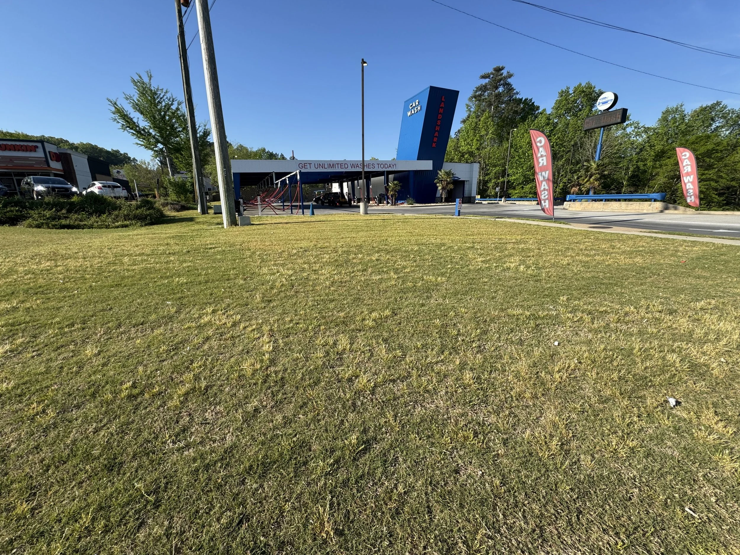 A freshly landscaped auto wash and detailing business with a large blue sign next to the building, advertising unlimited washes, with flags reading 'Wash' and 'Dry'. There are parked cars and trees in the background.