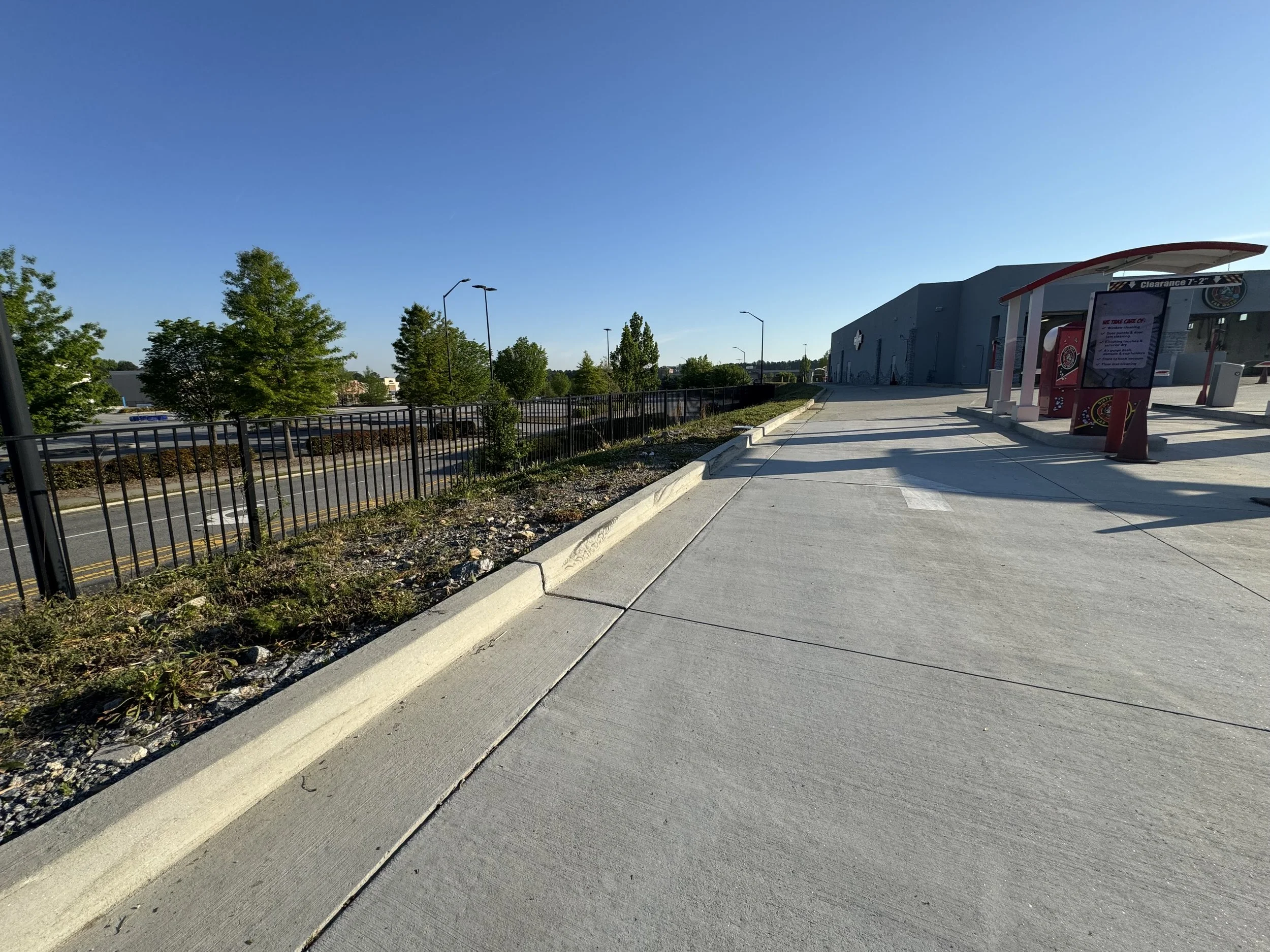 A sidewalk next to a building with a red and white overhang, a parking lot, and trees under a clear blue sky.
