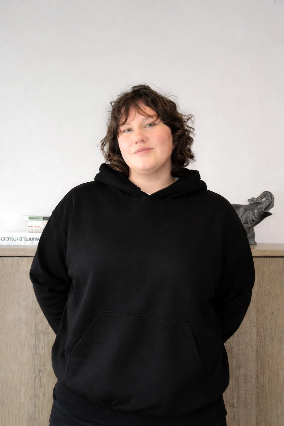 Junge Frau mit braunen, lockigen Haaren trägt schwarze Hoodie in einem Raum, steht vor einer Wand mit Holzwand. Im Hintergrund befinden sich Bücher und eine dekorative Statue.
