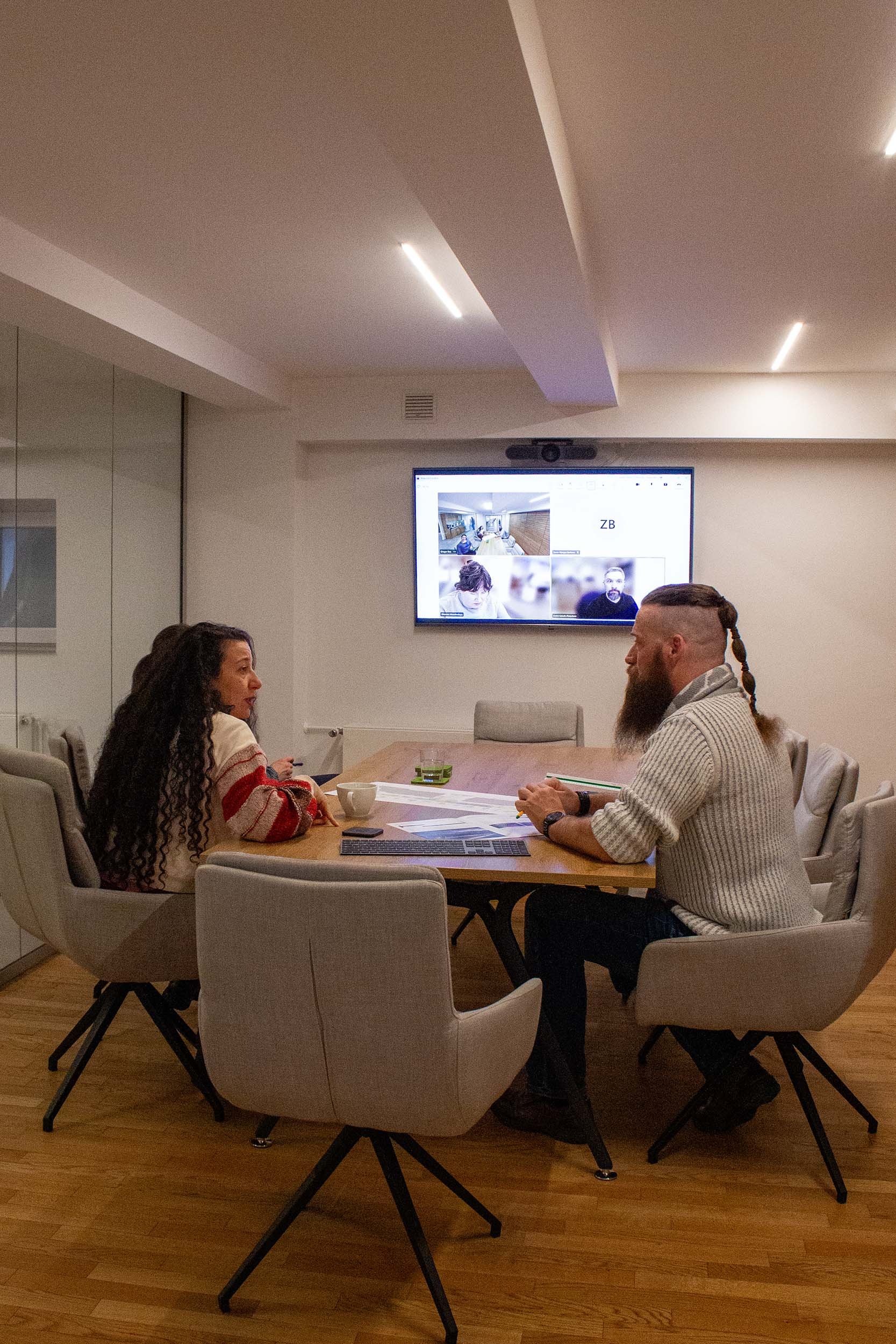 Zwei Personen sitzen an einem runden Tisch in einem Besprechungsraum, während auf einem Bildschirm im Hintergrund eine Video-Konferenz mit mehreren Teilnehmern läuft.