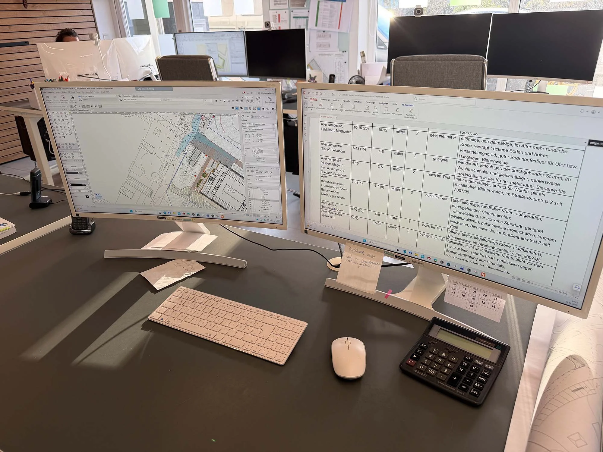 Zwei Computermonitoren an einem Schreibtisch, auf denen Baupläne und Tabellen zu sehen sind, umgeben von Bürobedarf wie Tastatur, Maus, Taschenrechner, Telefon und Notizen, in einem modernen Büro mit mehreren Arbeitsplätzen.