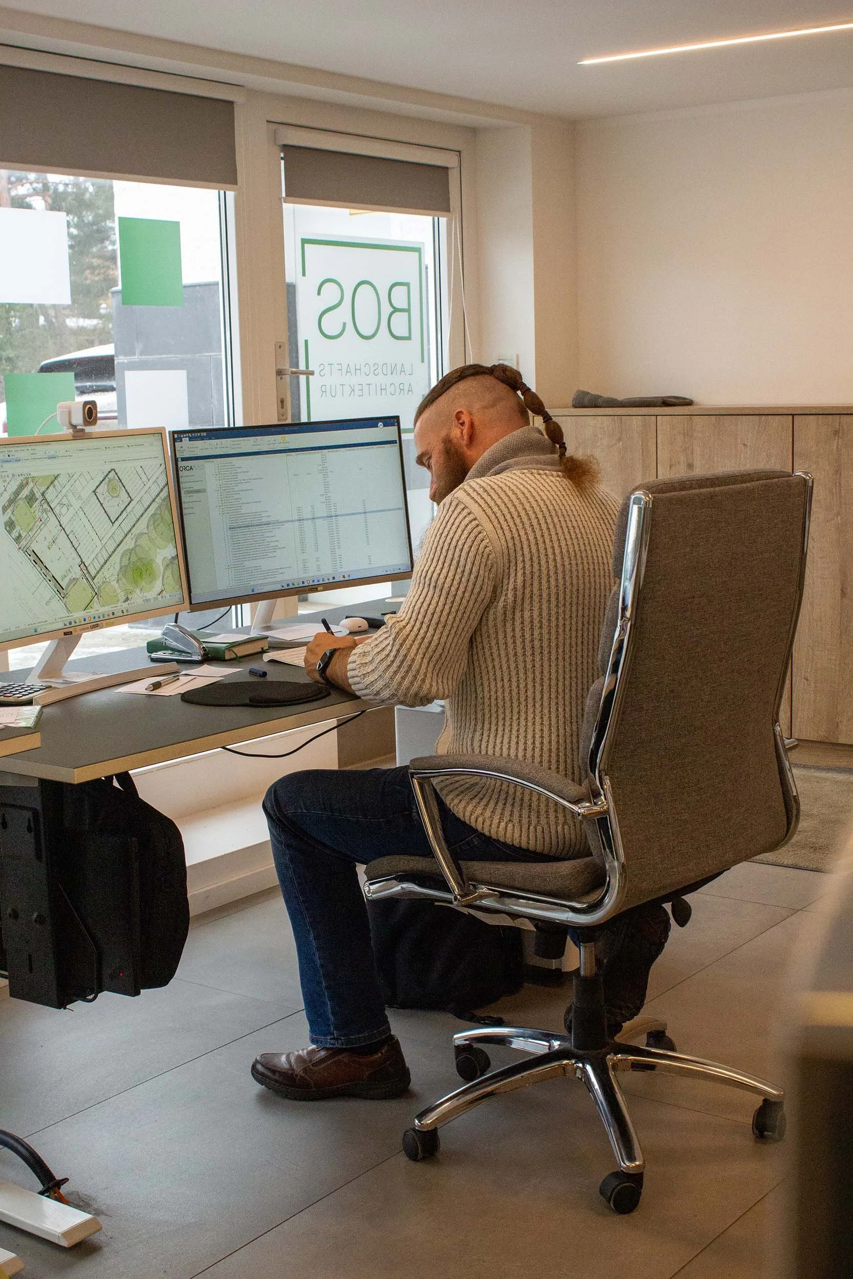 Ein Mann sitzt an einem Büroarbeitsplatz mit zwei Monitoren, arbeitet an seinem Schreibtisch und lächelt.