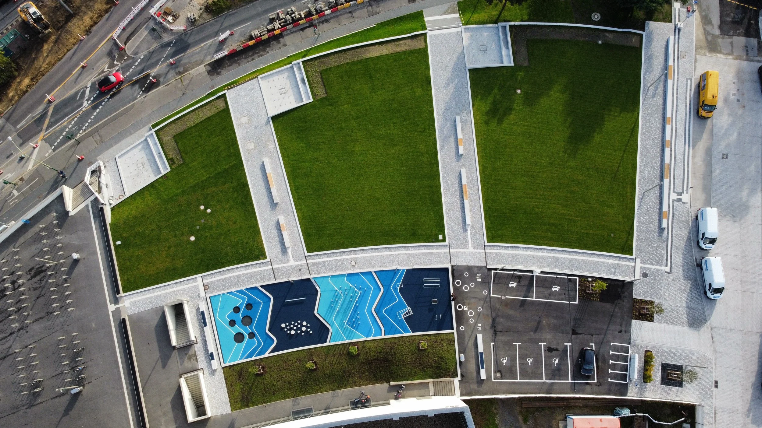 Luftaufnahme eines modernen Gebäudes mit grünen Dachflächen, einer Parkfläche und einem Farbfeld mit geometrischen Mustern.