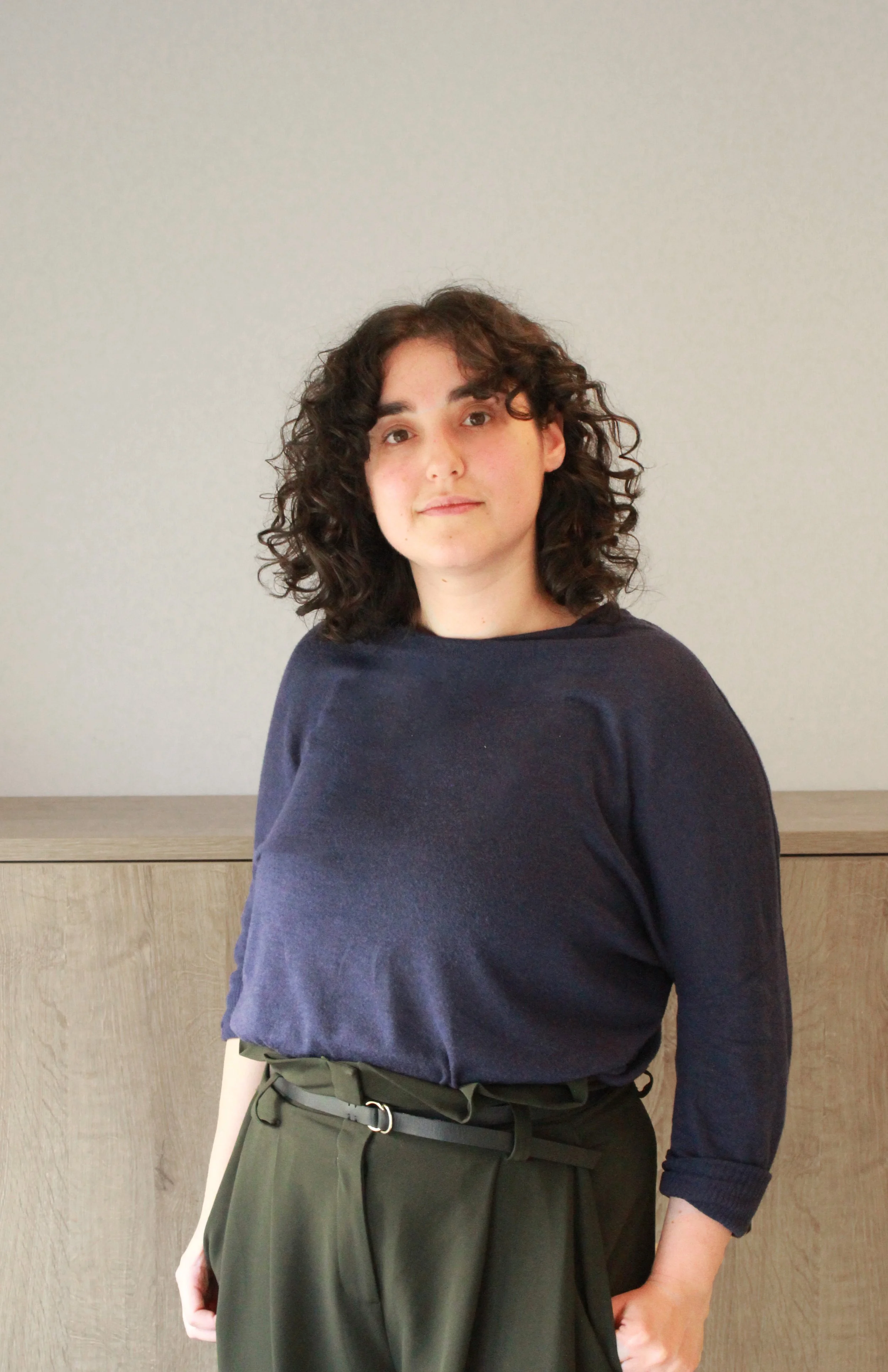 Junge Frau mit lockigem braunem Haar, dunkler blauer Pullover und grünen Hosen steht vor einer hellen Wand und schaut in die Kamera.