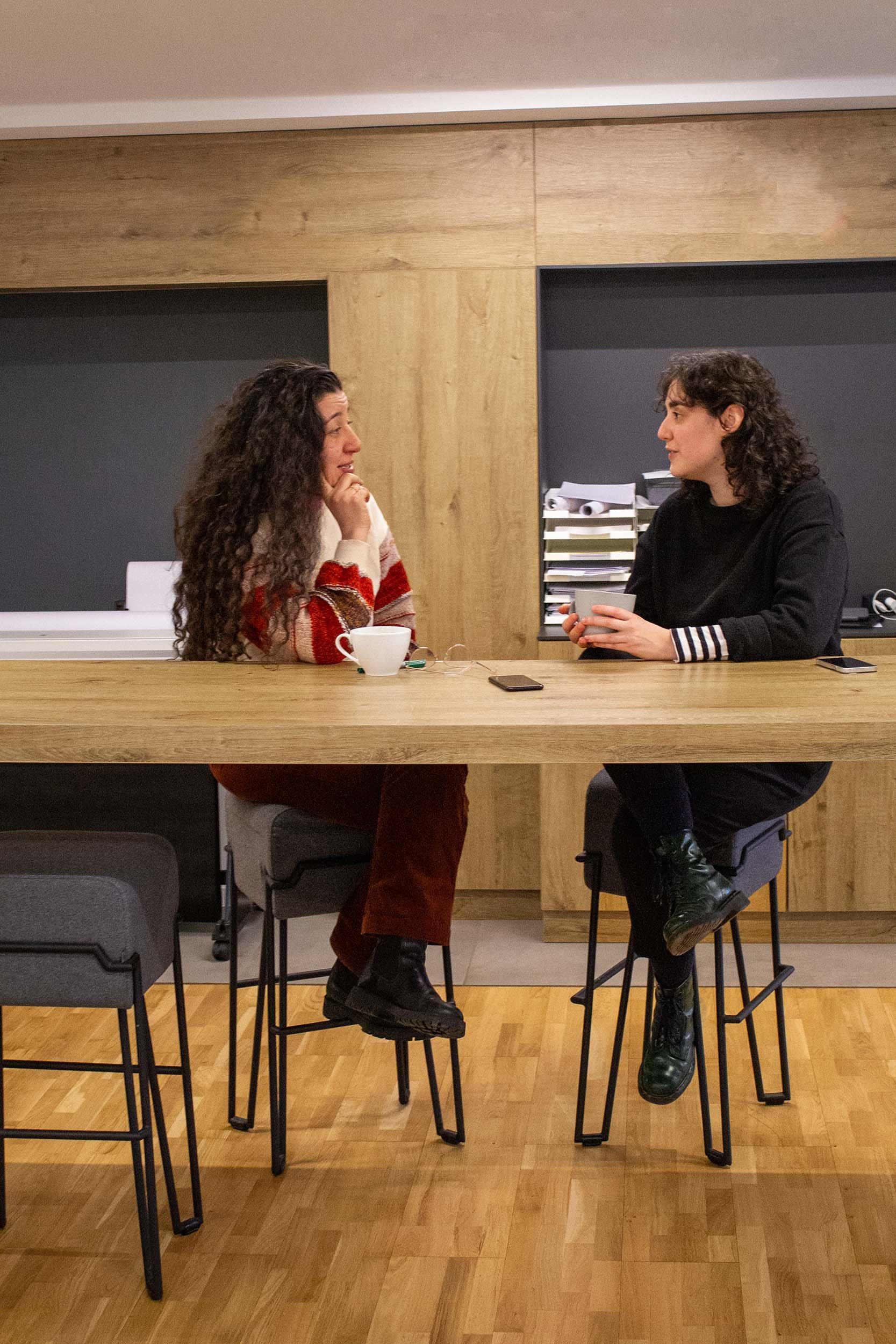 Zwei Frauen sitzen an einem Holztisch, unterhalten sich, trinken Kaffee, in einem modernen Raum mit Holzverkleidung.