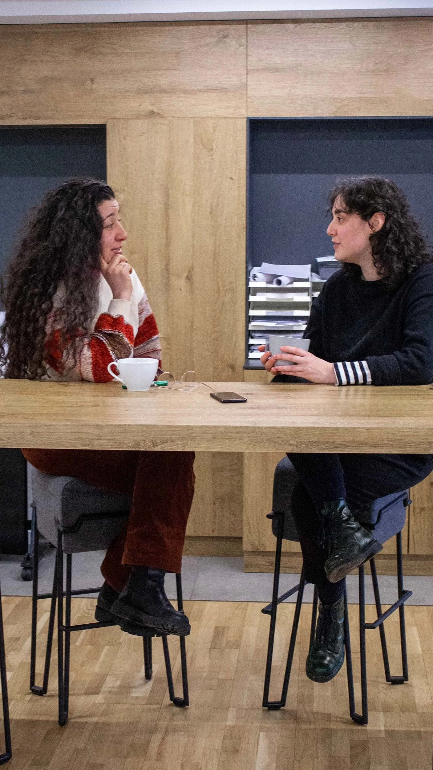 Zwei Frauen sitzen gegenüber an einem Holztisch, unterhalten sich und trinken Kaffee.