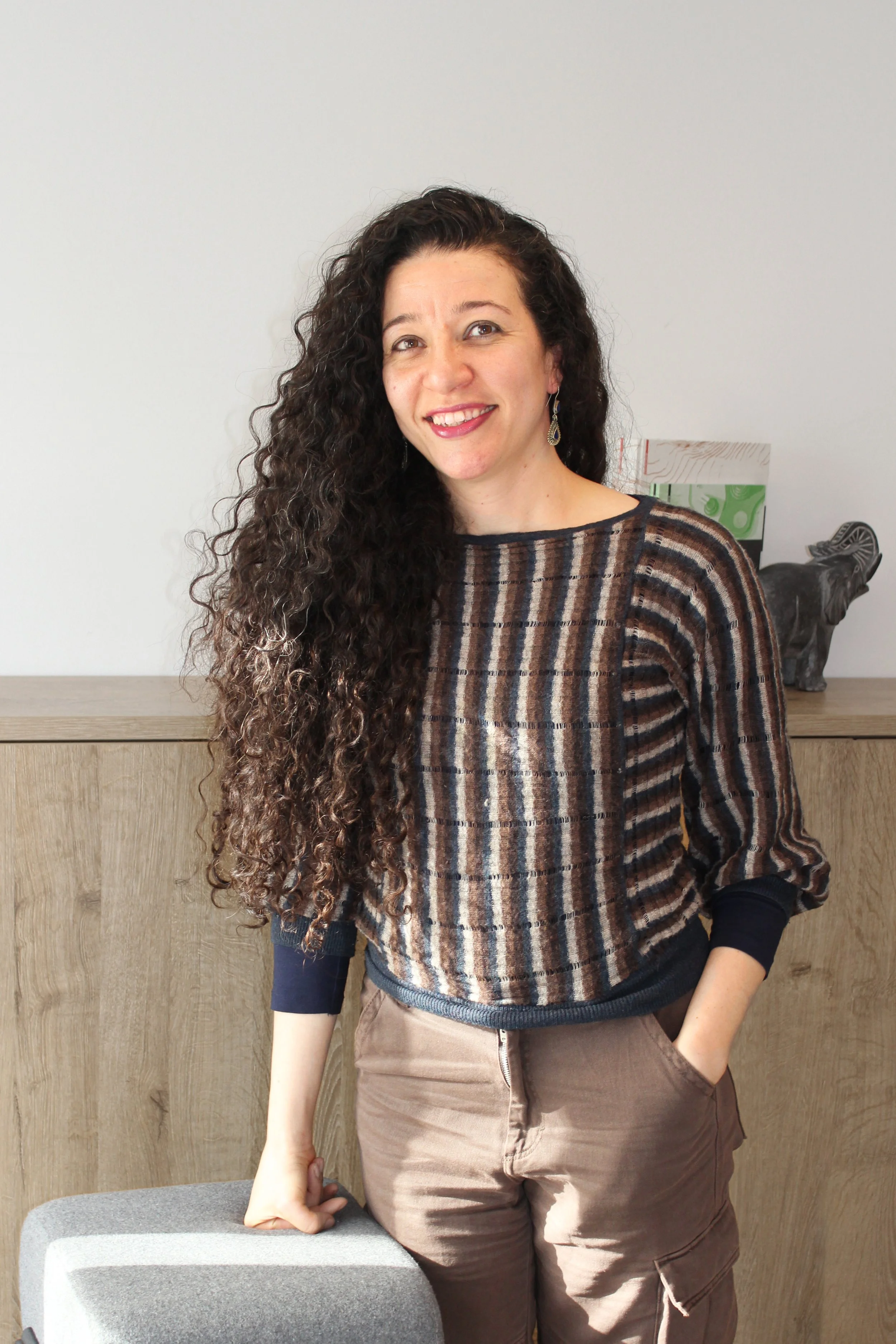 Eine Frau mit langen, lockigen dunklen Haaren, die lächelnd vor einer Holzwand steht, trägt ein gestreiftes oberteil und braune Hosen.