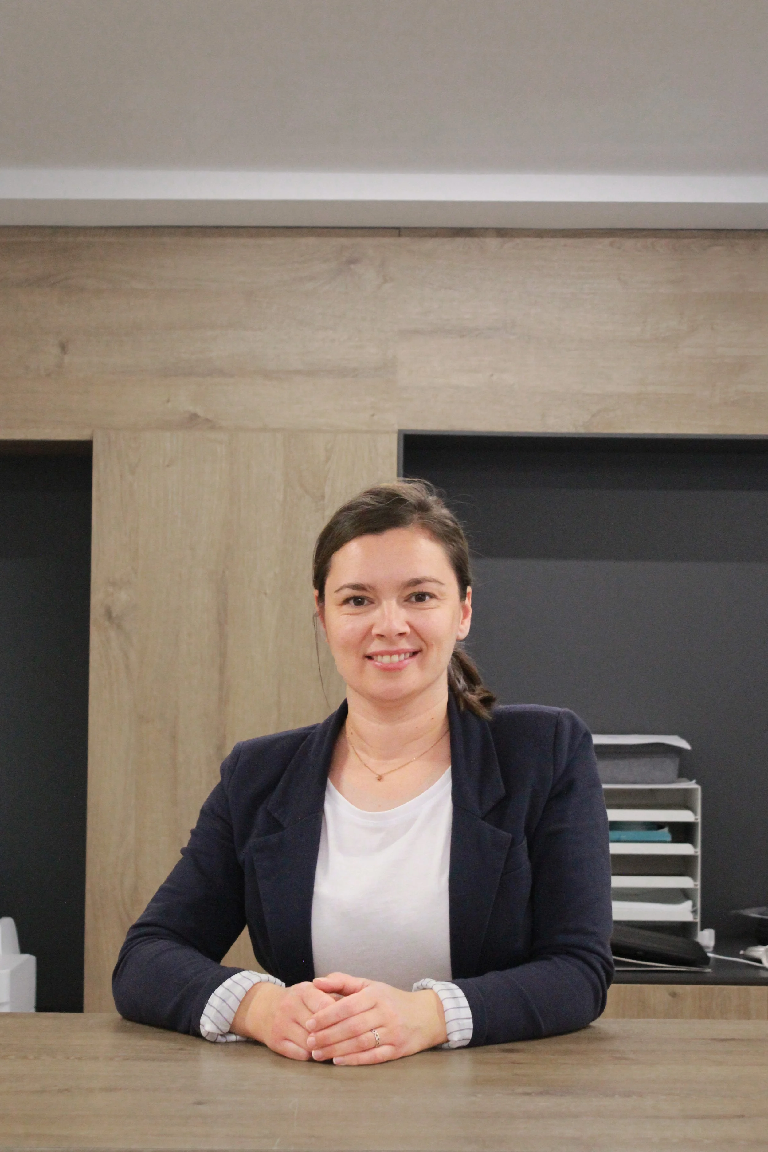 Eine Frau sitzt an einem Schreibtisch in einem modernen Büro. Sie trägt eine dunkle Jacke und ein weißes T-Shirt, lächelt, hat braune Haare, die zu einem Zopf gebunden sind, und blickt in die Kamera.