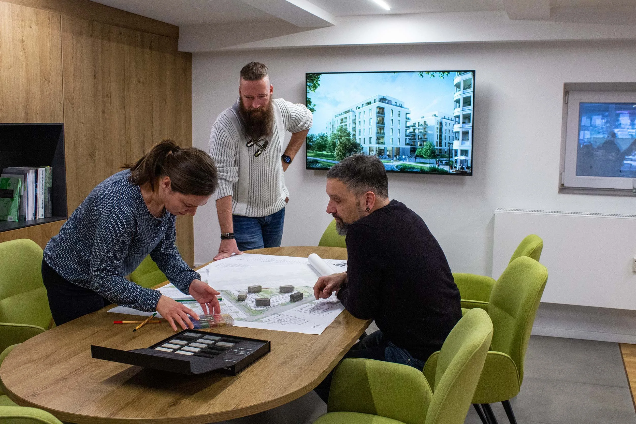 Drei Menschen diskutieren um einen Tisch mit Bauplänen und Mustern. Im Hintergrund ein Bildschirm mit einer Wohnanlage, im Raum sind grüne Stühle.