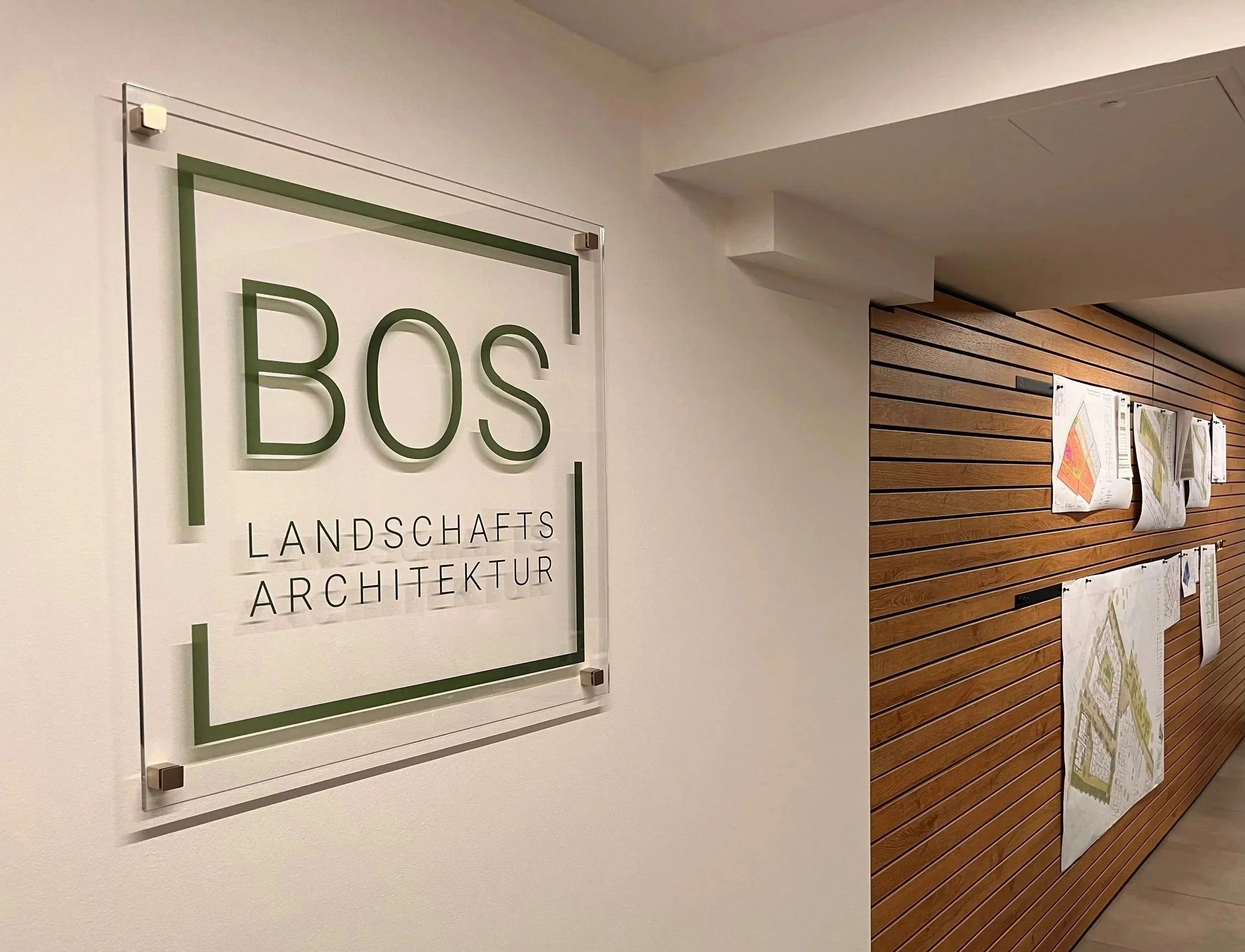 Innenansicht eines Büros mit einem großen Schild an der Wand, das 'BOS Landschafts Architektur' heißt, neben einer Holzpaneelwand, an der Pläne und Karten hängen.