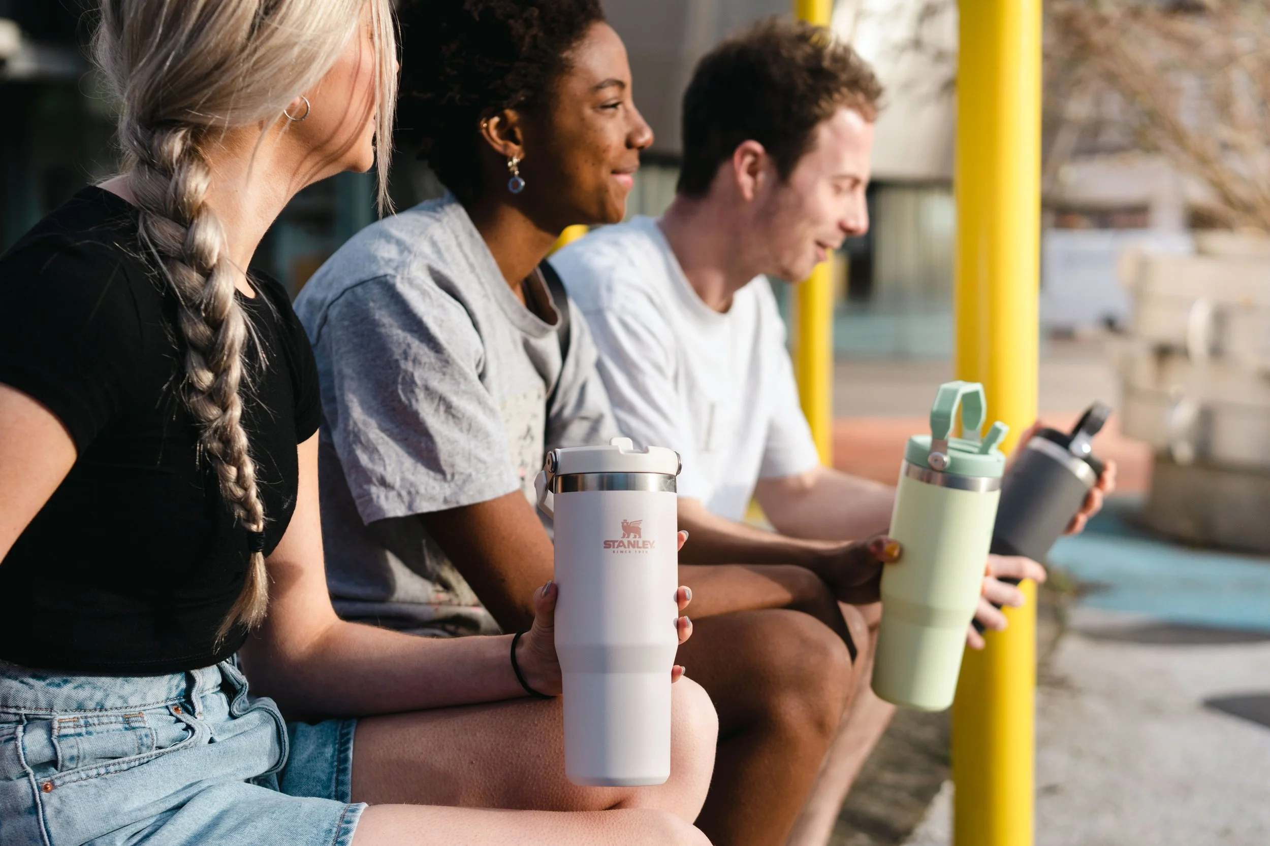 Three young people sitting on a bench outdoors, holding Yeti travel mugs and looking at their phones, with an athletic corporate shirt on.