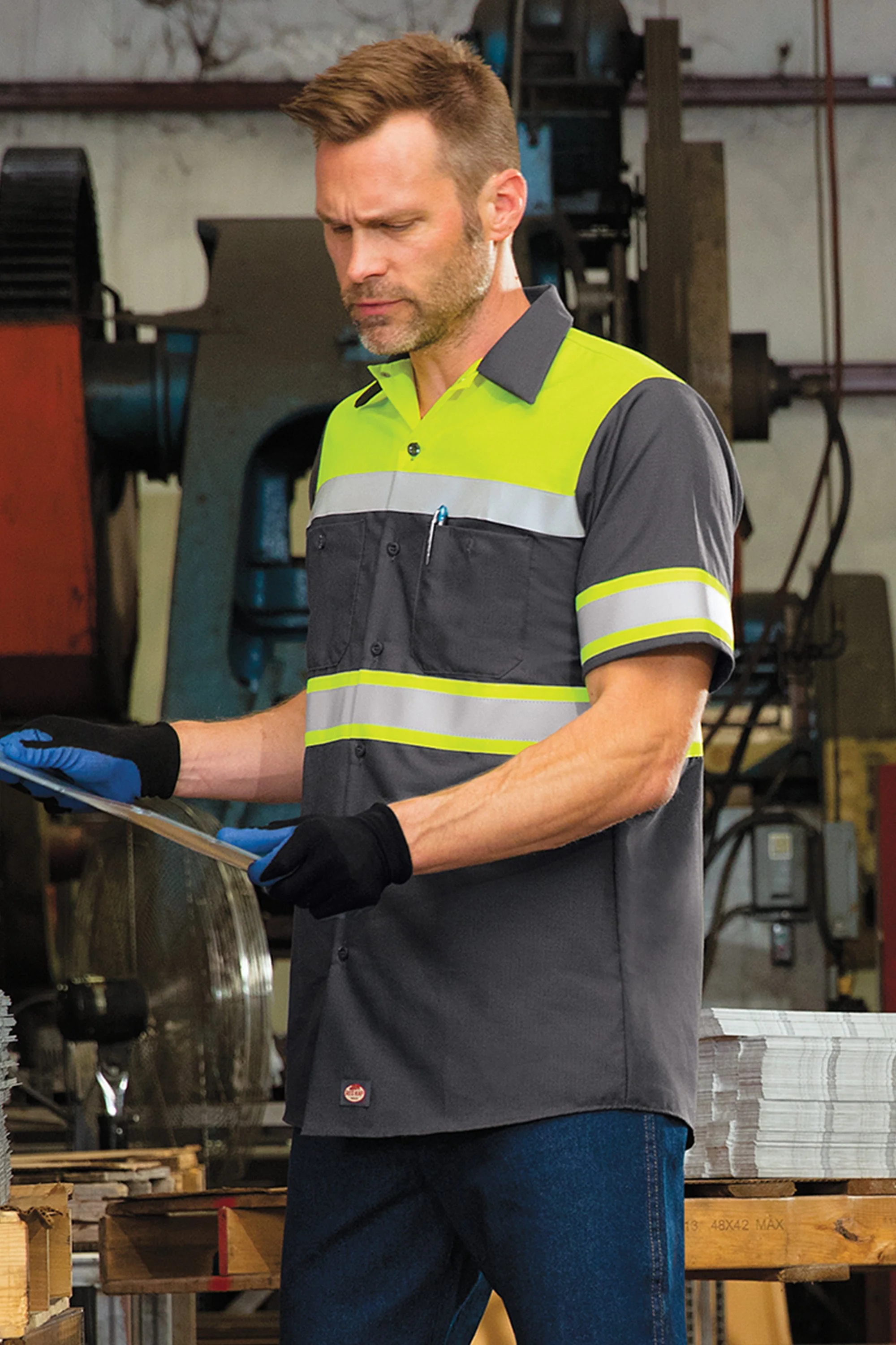A man wearing a gray and neon yellow safety shirt and black gloves inspecting documents in an industrial warehouse.