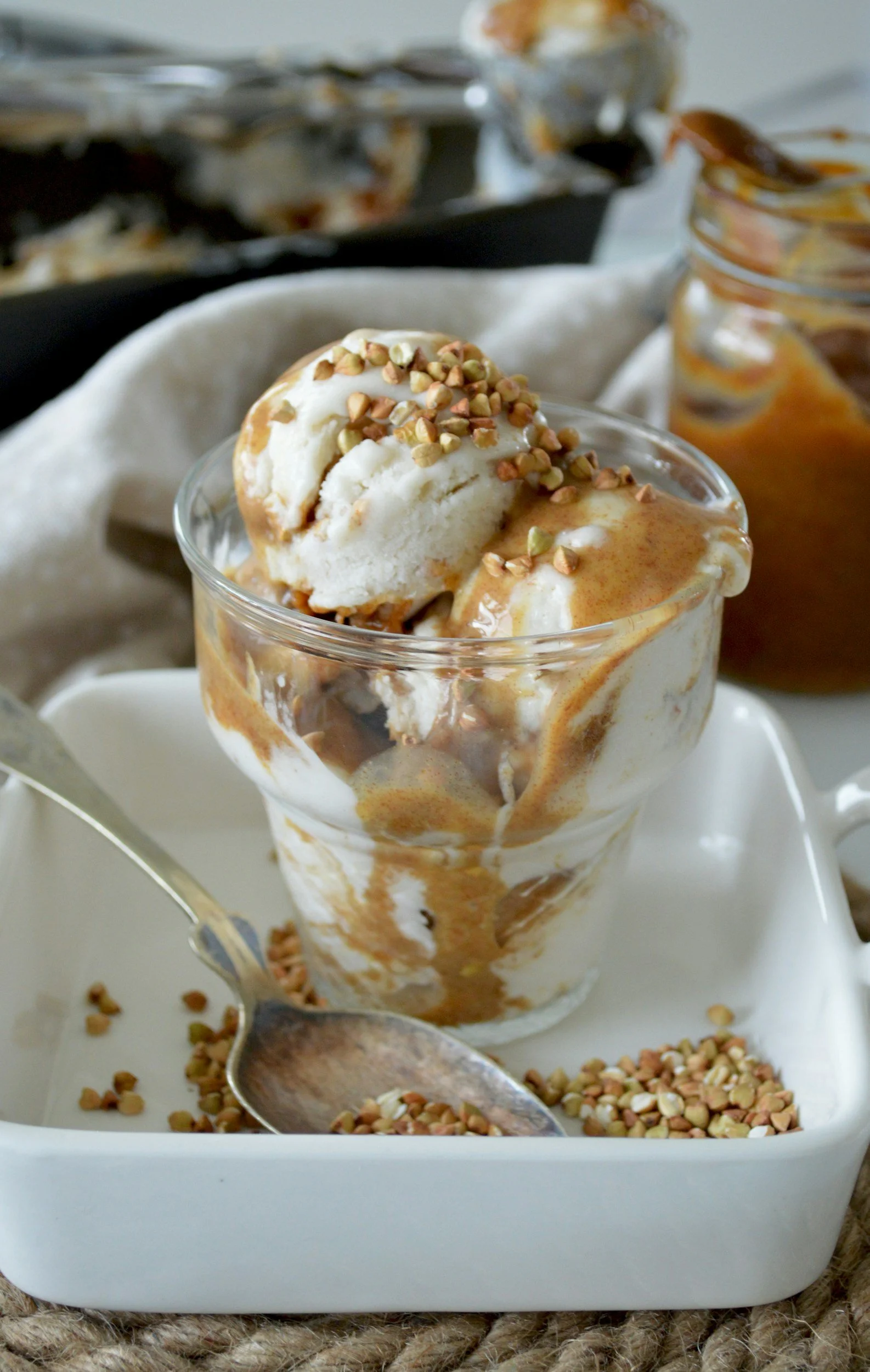 A glass of ice cream topped with chopped nuts and caramel sauce on a white tray, with a spoon inside and additional nuts scattered on the tray.