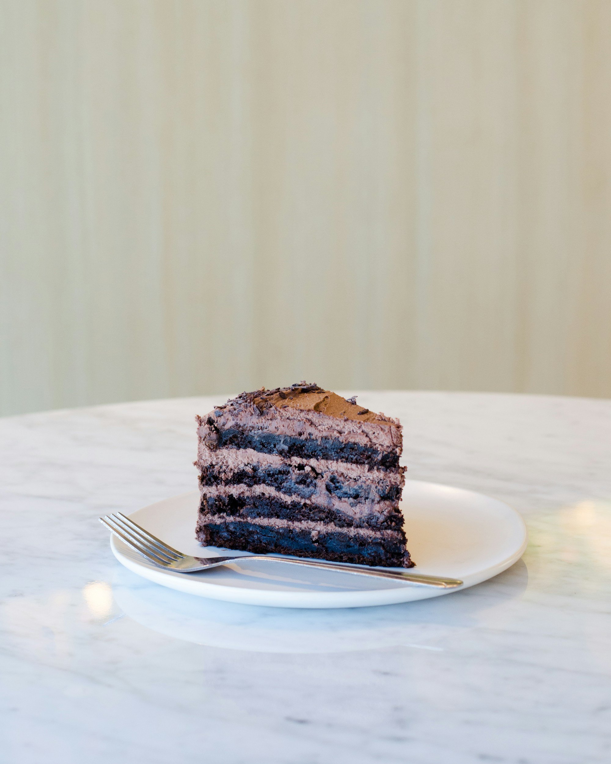 A slice of layered chocolate cake on a white plate with a fork, on a marble table with a plain beige wall in the background.