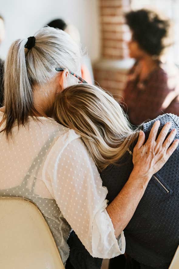 An elderly woman with gray hair and glasses hugs a younger woman, resting her head on her shoulder in a comforting gesture in a cozy room.