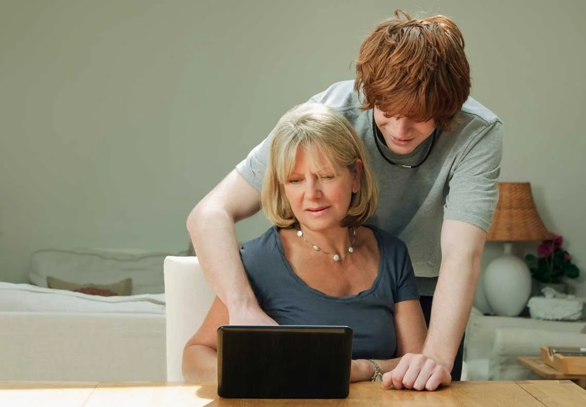 A young man is helping an older woman look at a tablet together in a living room.