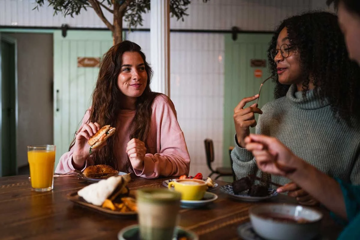 Two women sitting at a wooden table having breakfast, with plates of food, glasses of orange juice, and a bowl of syrup, engaging in conversation.