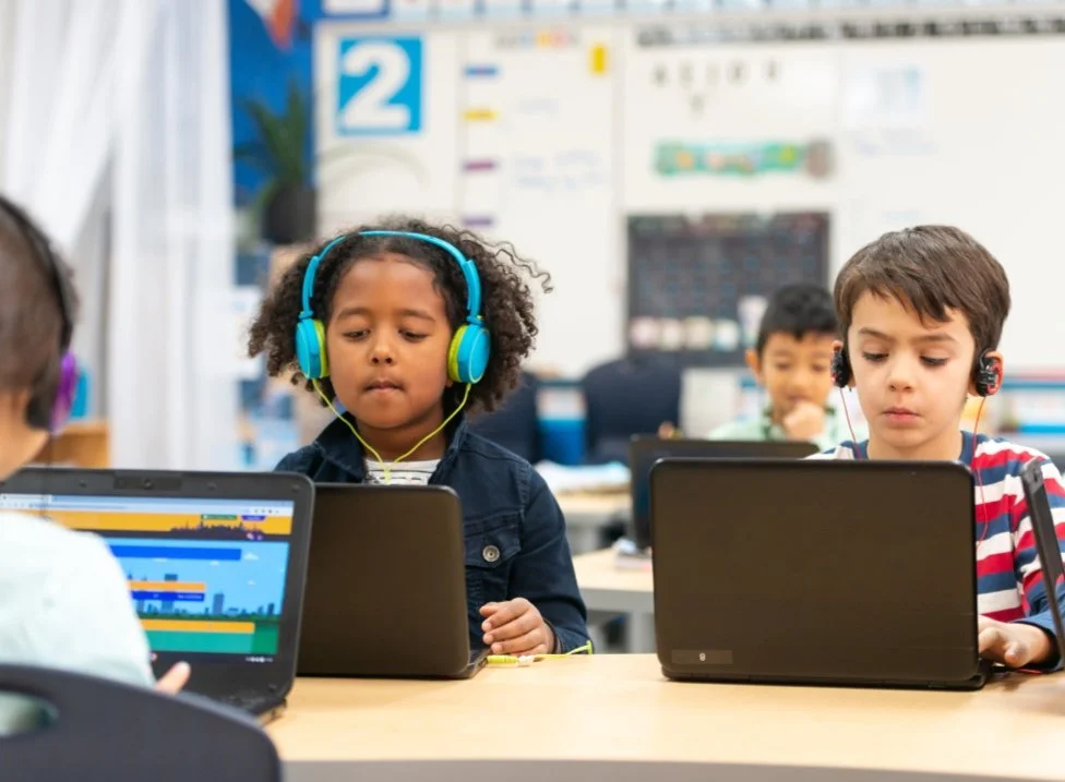 Children wearing headphones using laptops in a classroom setting.