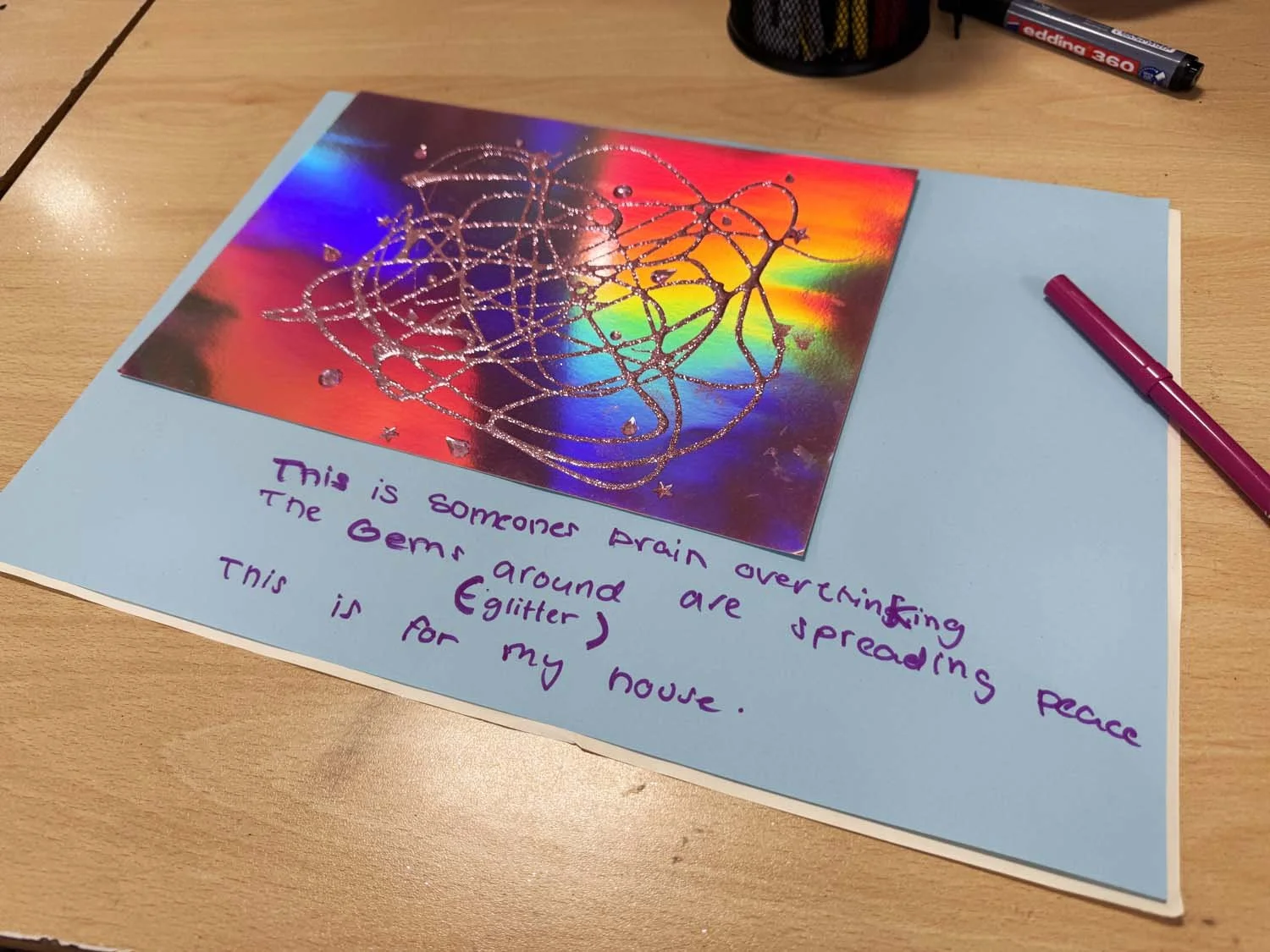 A child's drawing with glue and glitter on a holographic panel attached to blue paper, featuring handwritten text that reads, "This is someone's brain overthinking."