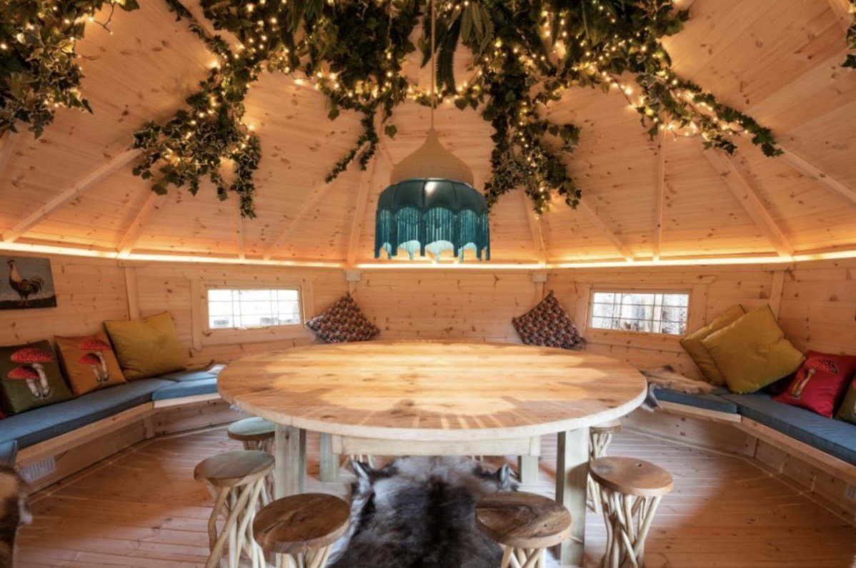 Interior of a cozy wooden roundhouse with a central round table, rustic stools, and bench seating with colorful cushions. The ceiling features green foliage and string lights, with a blue pendant lamp hanging above.