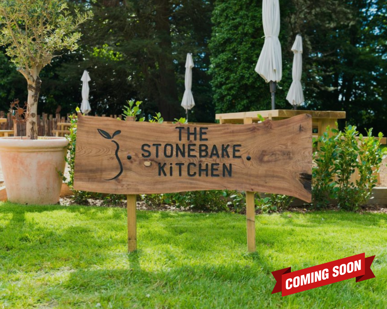 Wooden sign reading 'The Stonebake Kitchen' in outdoor garden setting with potted plants and closed umbrellas.