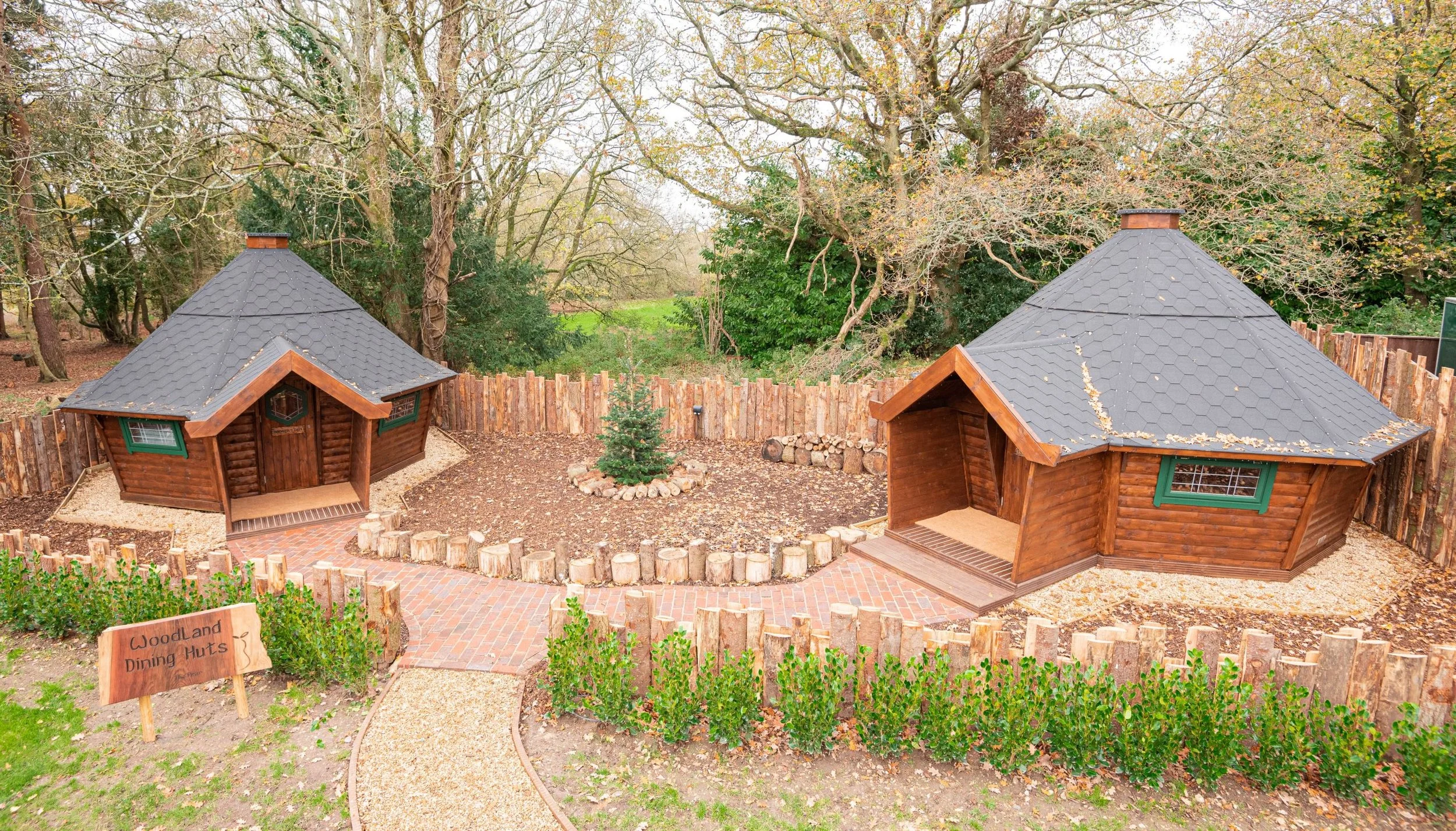 Two wooden hexagonal huts with shingled roofs in a forest setting, surrounded by a fenced area with paths, greenery, and a central small pine tree. A "Woodland Dining Huts" sign is visible in the foreground.