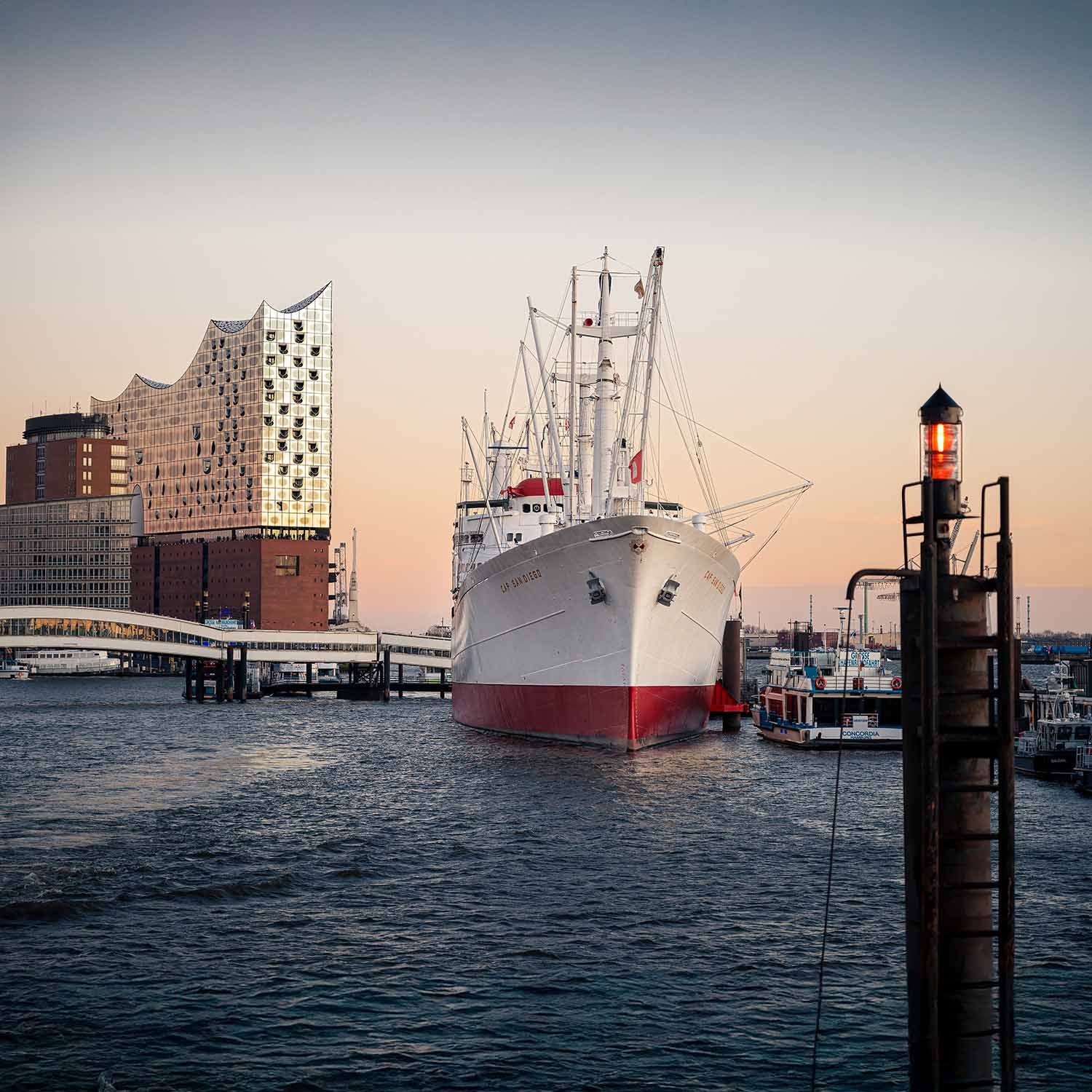 Large white and red ship docked at a harbor with modern buildings in the background during sunset.